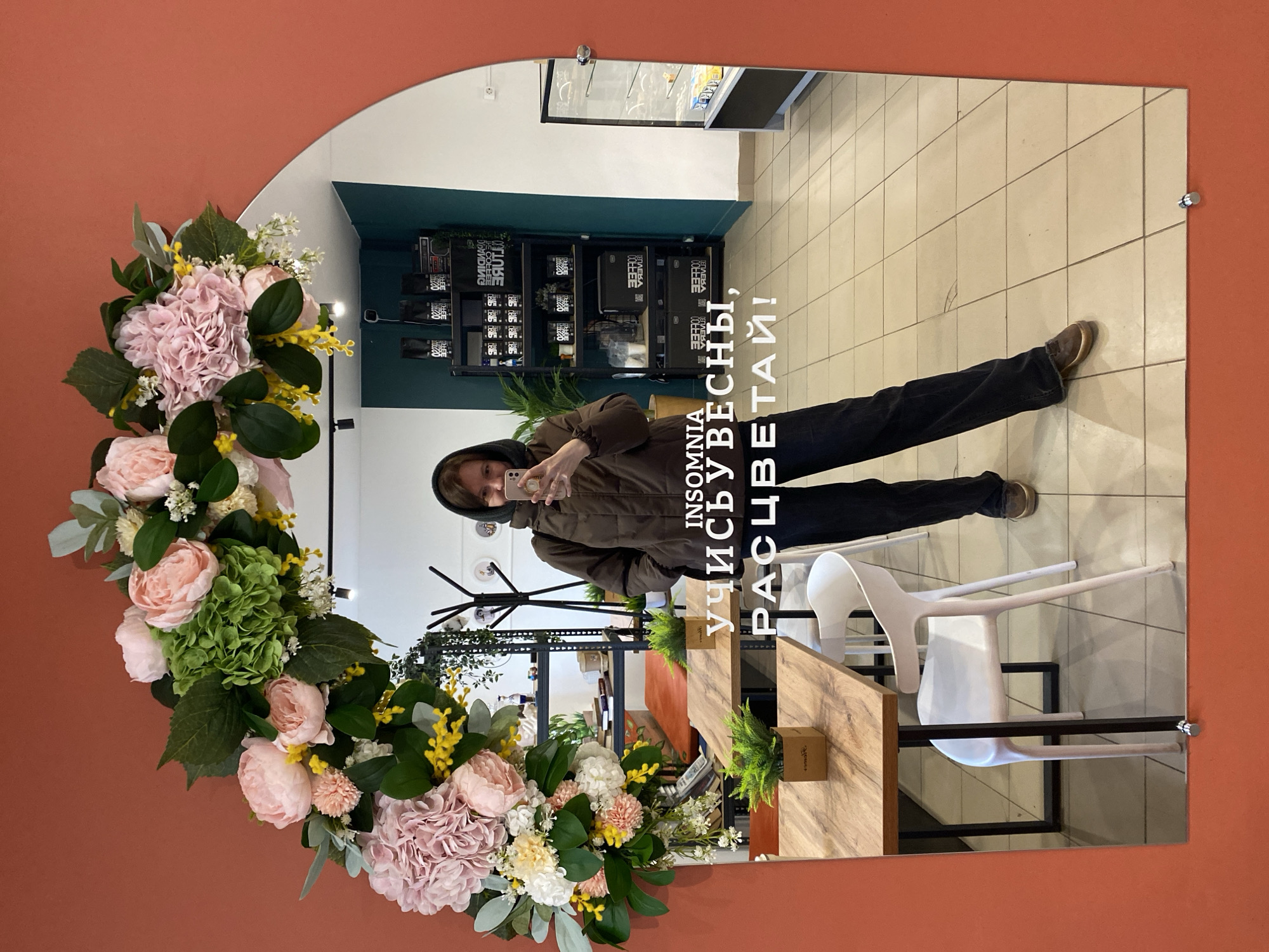 A person in a brown hooded jacket and dark pants stands in front of an arched mirror taking a selfie. The mirror is decorated with a wreath of artificial flowers. The reflection shows a cafe interior with tables, chairs, a vending machine, and retail displays. The cafe appears to be in Chaikovsky, Russia, based on the Cyrillic lettering "INSOMNIA, УЧИСЬВЕСНЫ, РАСЦВЕТАЙ!" visible in the mirror. The flooring is light-colored tile.