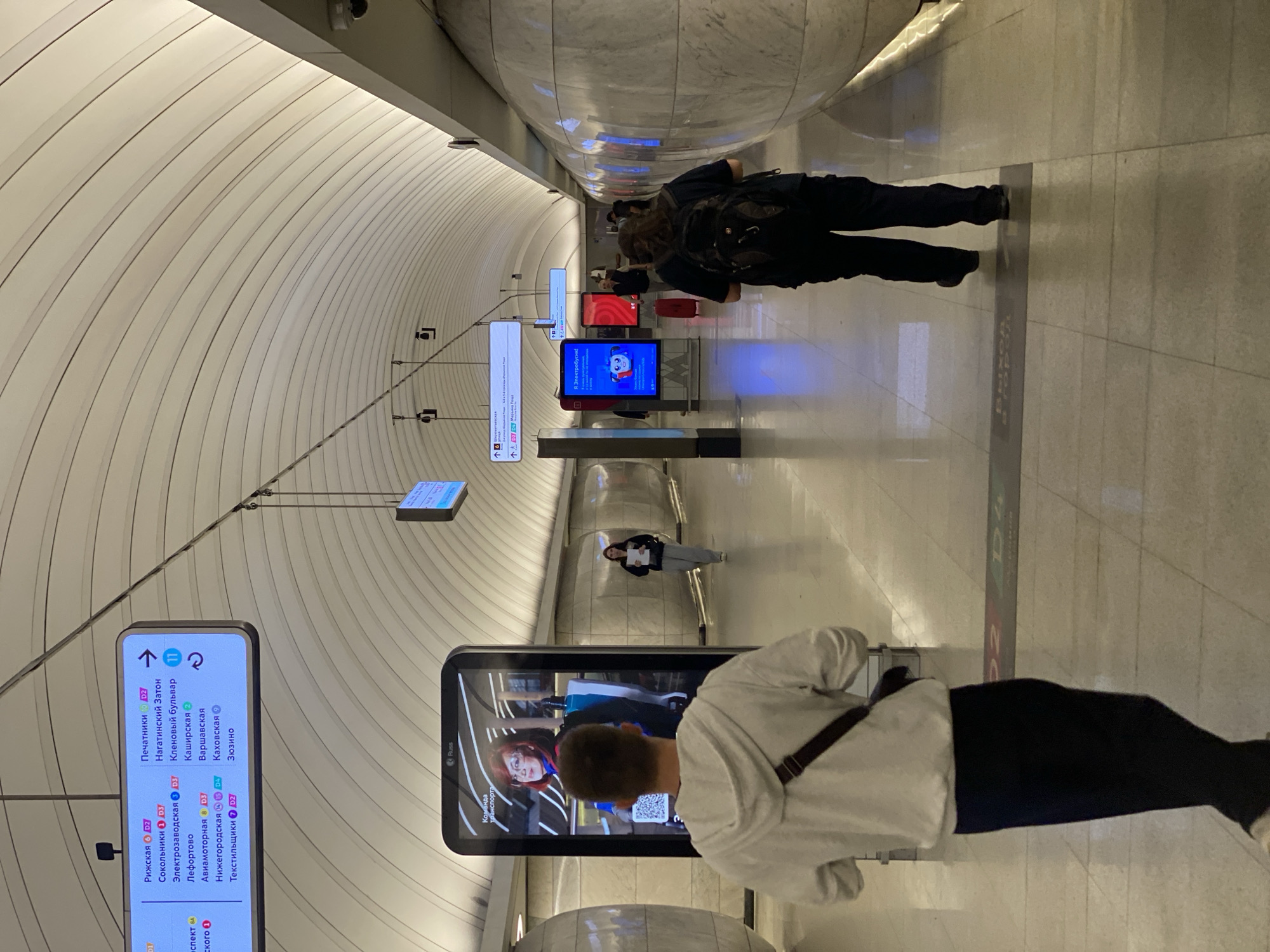The image depicts a Moscow Metro station platform. In the foreground, a person wearing a white hooded sweatshirt and dark pants walks away from the camera, carrying a dark-colored satchel across their chest. To the left, a large digital advertisement displays a woman with red hair. Further down the platform, another person in dark clothing with a backpack walks away from the camera. Several other individuals are visible in the distance, also walking along the platform. Overhead, electronic signs display station names and line information in Russian, including "Рижская" (Rizhskaya), "Сокольники" (Sokolniki), and "Печатники" (Pechatniki), indicating this is likely a station on the Sokolnicheskaya (Red Line) or Lyublinsko-Dmitrovskaya (Orange Line) of the Moscow Metro. The station features a distinctive curved ceiling with recessed lighting and marble-clad walls.