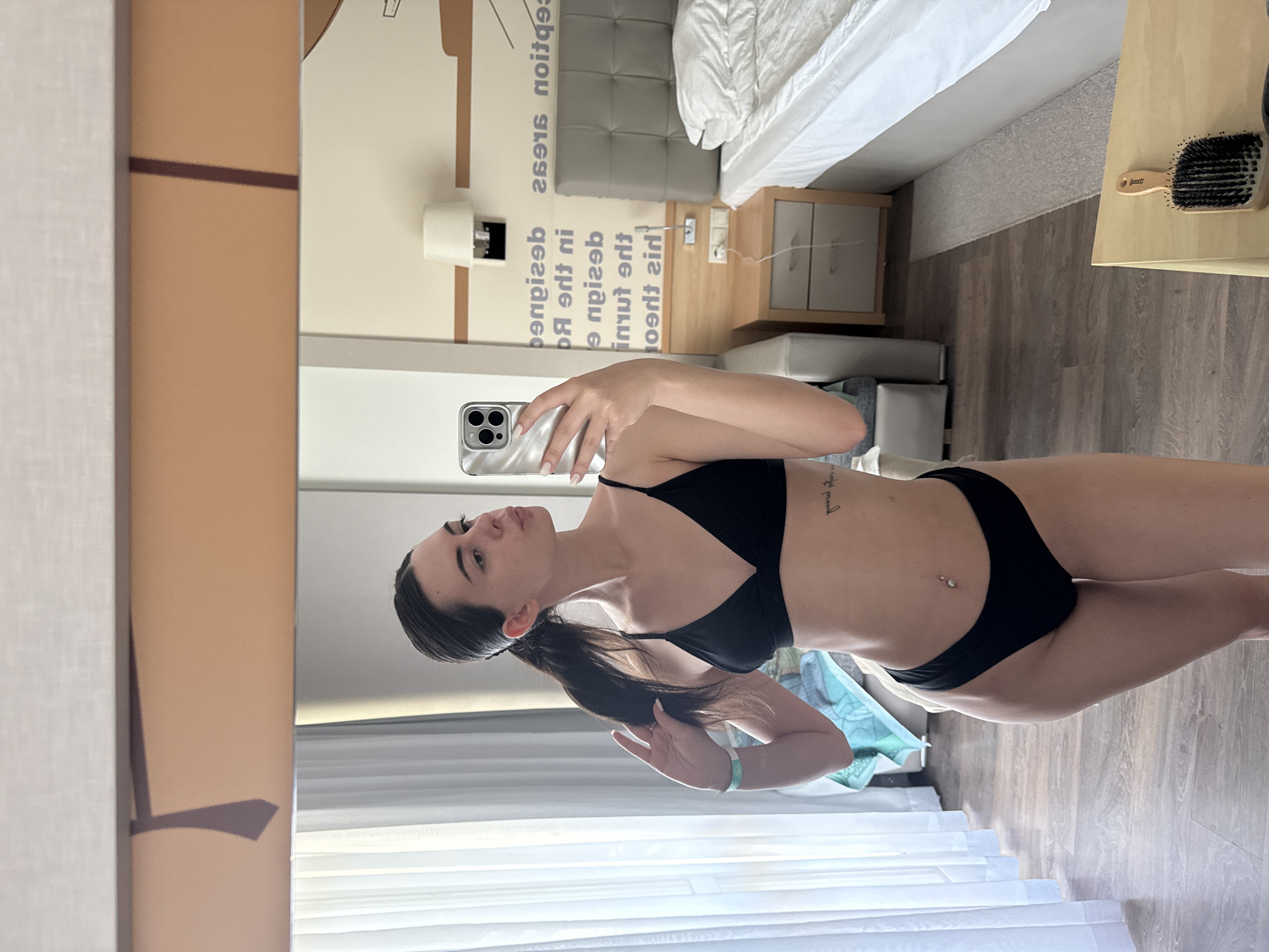 A young woman with dark hair pulled into a ponytail stands in a hotel room, taking a mirror selfie. She is wearing a black bikini. Her left hand is raised, touching her hair. A portion of a white towel is visible on the floor behind her. The room features a wooden floor, a bedside table with drawers, a lamp on the wall, and a bed with white bedding. The wall behind the bed has a textured design with text. A window with white curtains is visible to the left of the woman.