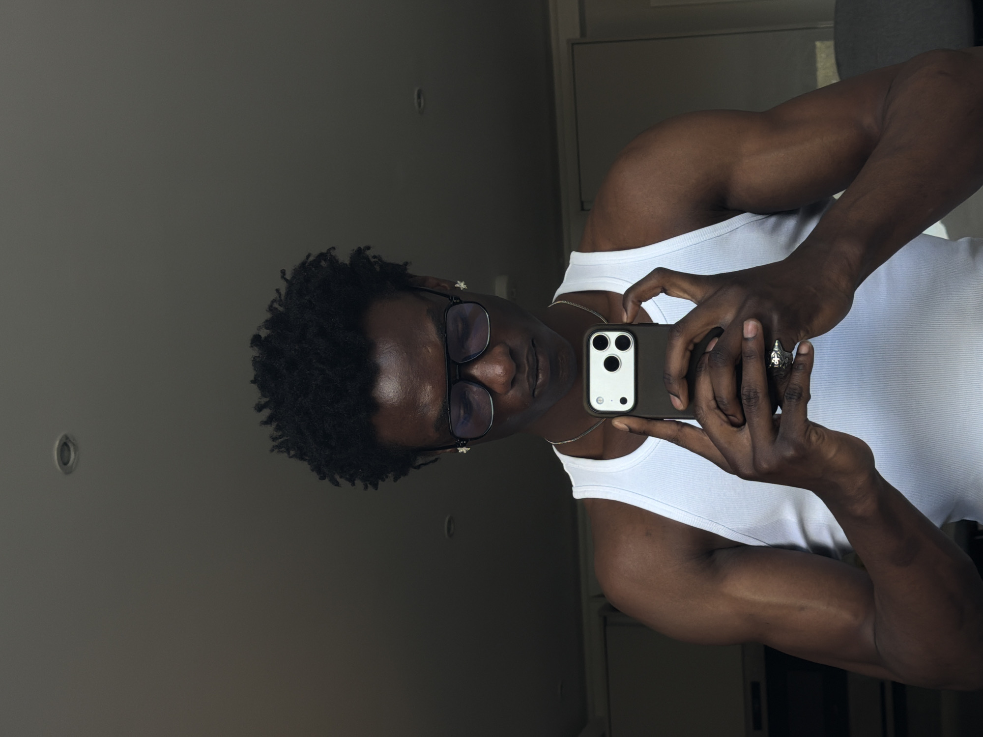 A dark-skinned man is captured in a mirror selfie, holding a smartphone to photograph his reflection in an indoor setting. He is positioned vertically, looking towards the camera with a neutral expression. He has short, dark, curly hair and is wearing a white ribbed tank top. Clear-framed glasses with dark lenses cover his eyes, and a thin silver chain necklace adorns his neck. On his right ring finger, he wears a prominent silver ring with an intricate design. His arms appear muscular.
The man holds a dark-colored smartphone, identifiable by its triple camera lens system, with both hands, steadying it for the shot. The background reflected in the mirror is a plain, dark grey or olive-grey wall. A small, circular fixture or detail is visible on the wall to his left. The lighting is diffused, indicating an indoor environment, likely a room within a residential building. No explicit text is visible in the image.