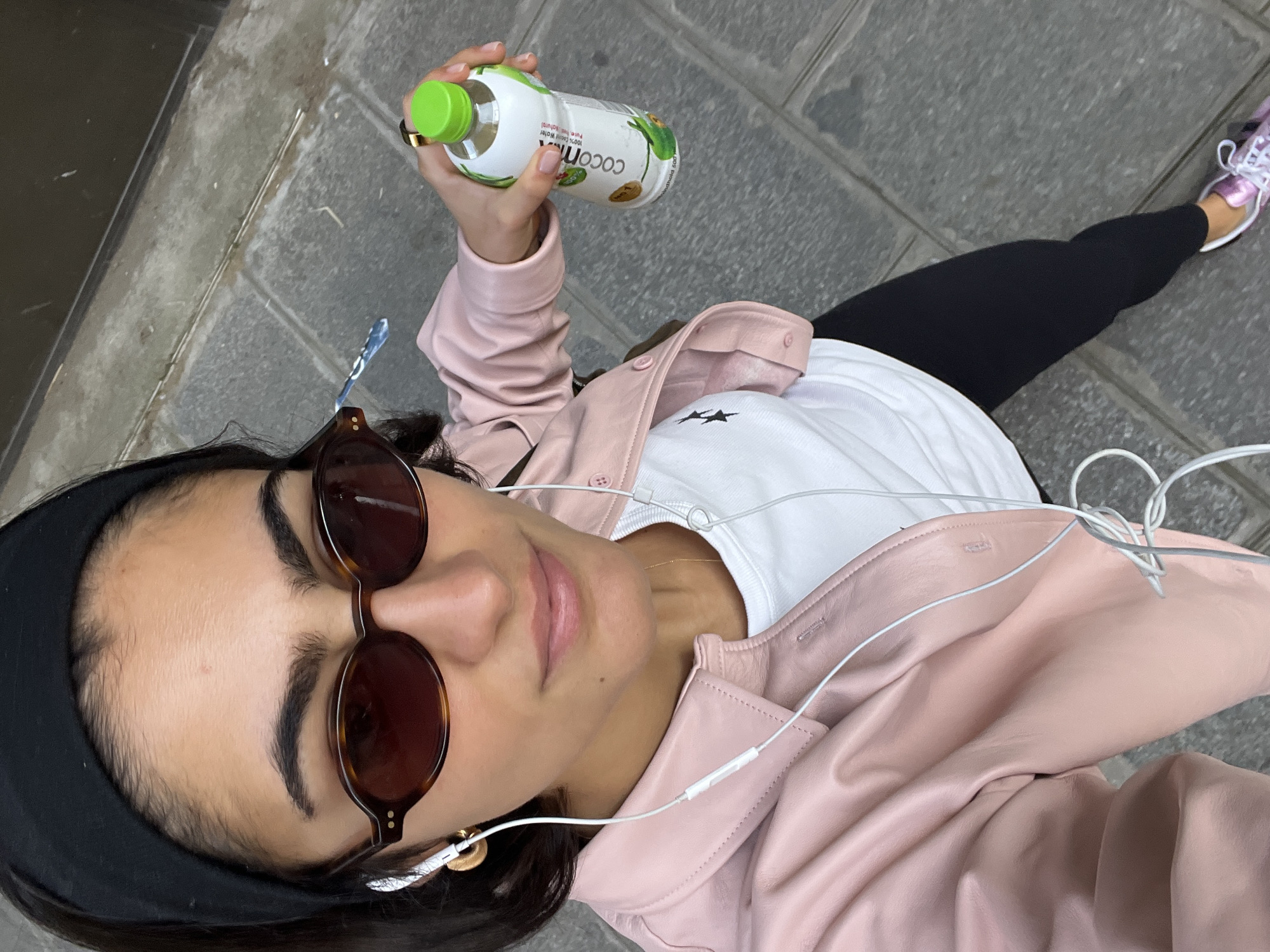 A woman in a pink leather jacket and sunglasses is holding a bottle of coconut water on a stone-paved street. She is wearing black leggings, a white shirt with a black star, and white headphones. She has a black headband in her dark hair. The scene appears to be in Paris, France, based on the architecture and street design.