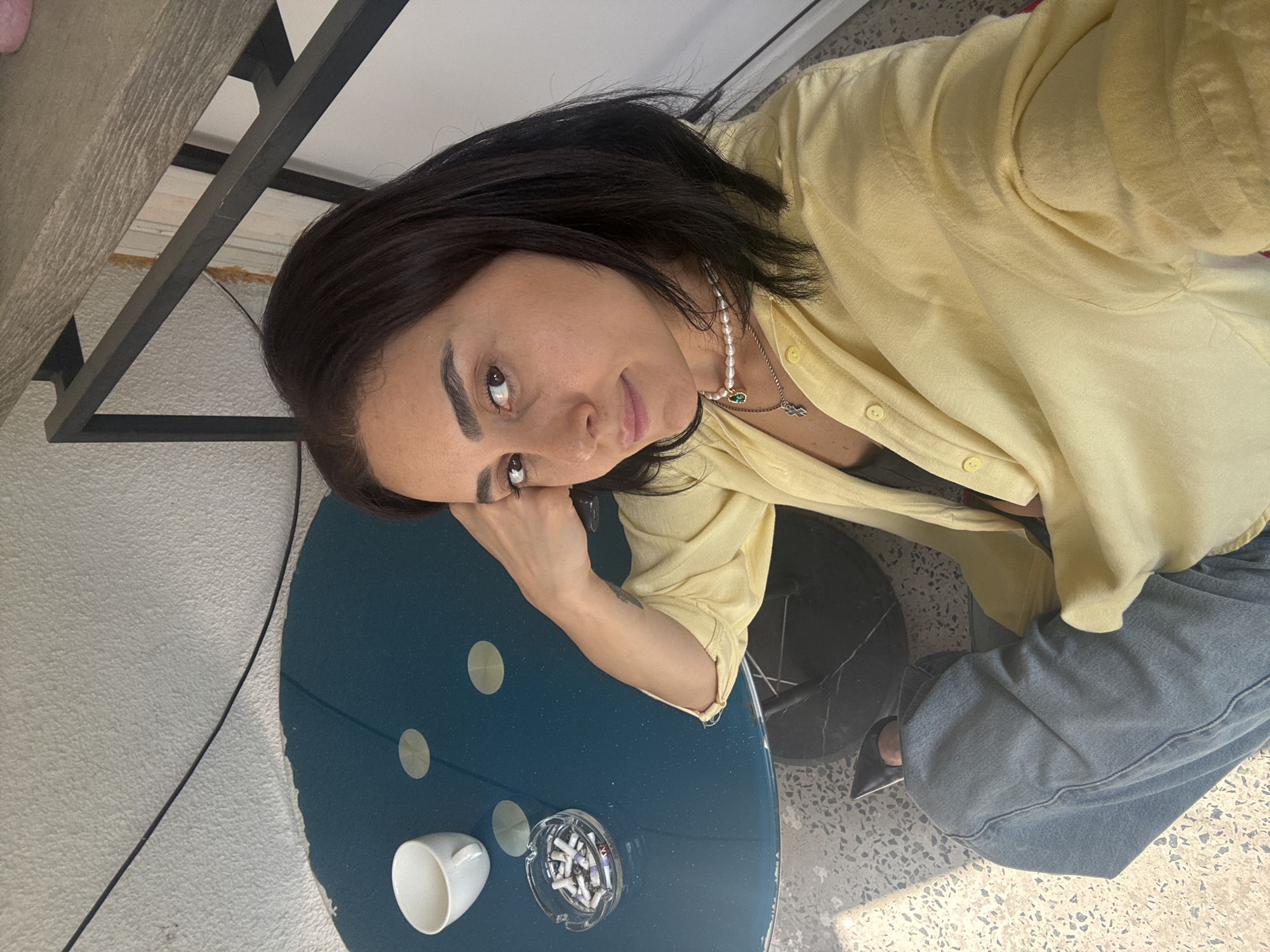 In a selfie taken in Tbilisi, Georgia, a woman with dark hair and brown eyes poses with her head resting on her hand. She is wearing a light yellow, button-down shirt and grey wide-leg pants. A pearl necklace with a green pendant adorns her neck.
The setting appears to be indoors, near a window, with natural light illuminating the scene, suggesting it could be daytime. A blue circular table is in the foreground, on which rests a white cup and an ashtray filled with cigarette butts. Behind her, a dark metal structure, possibly part of a table or shelf, is visible. The background shows a textured wall and a floor with a terrazzo-like pattern. The woman's expression is thoughtful, perhaps a little melancholic, as she gazes directly at the camera. The overall impression is one of a quiet, introspective moment.