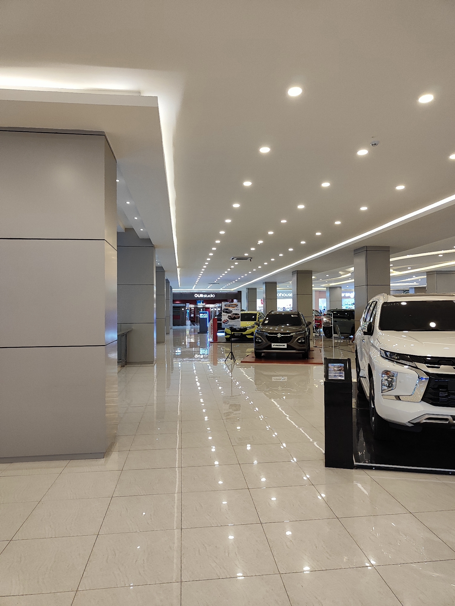 The image depicts the interior of a car dealership in Sempusari, Indonesia, likely located within a shopping mall given the polished tiled floor, abundant ceiling lights, and the presence of multiple cars on display. In the foreground on the right, a white SUV is prominently featured. Further into the dealership, several other vehicles are parked and displayed. A sign reading "OUR studio" is visible in the background, suggesting a branded display area or a partner company. The environment is well-lit and appears clean, with reflections on the floor indicating a highly polished surface. No people are visible in the image.