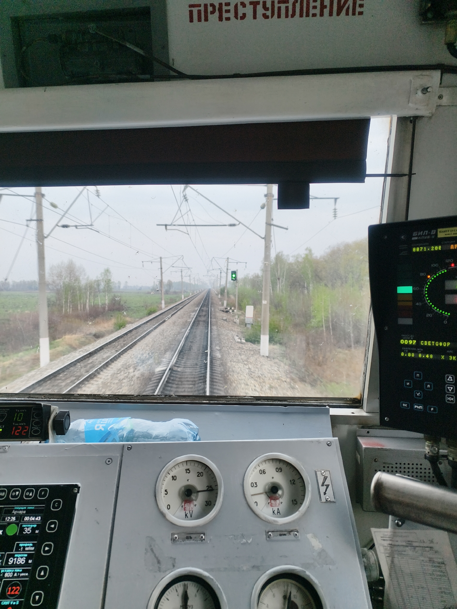 This image captures the view from inside the cabin of a train, traveling along a track in a rural environment, likely in Russia based on the Russian script.
**Environment:** The landscape is characterized by fields and trees on both sides of the railway. The weather appears overcast and the visibility is moderately clear. A green traffic light is visible along the track ahead. The track itself is dual-rail.
**Objects:**
* The train cabin features various control panels with dials and displays showing data like voltage (kV), current (kA), speed (in km/h), and other operational parameters. The dials are in Cyrillic script.
* A black box-like electronic device with a screen shows the traffic light status.
* A water bottle is seen on the control panel.
**Inscriptions:**
* The inscription "ПРЕСТУПЛЕНИЕ" (Prestupleniye) is visible above the window.
* Dials with "KV" and "KA" are visible, indicating voltage and current.
**Location:** Based on the signs, it is likely Russia or another former Soviet state. The city cannot be determined from the image, because there is no city in the image.