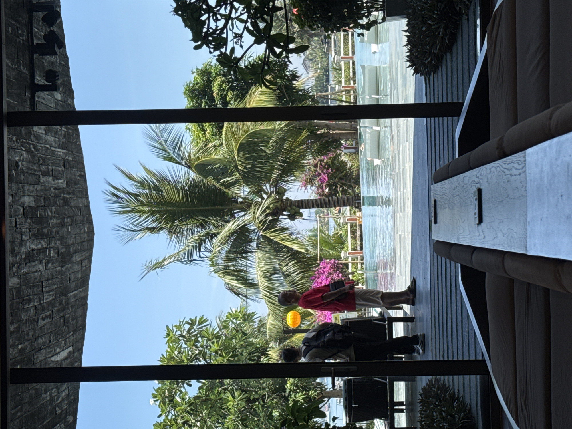 The image, taken in Đà Nẵng, Vietnam, shows an outdoor scene viewed from an interior location. A palm tree with coconuts is centered in the view, standing between a low building and a pool. In the foreground, two people are visible. One is wearing a red shirt and beige shorts, and the other has a backpack. They are standing on a wooden deck. The interior location has a dark stone wall on one side and an interior space with a cushioned bench on the other.