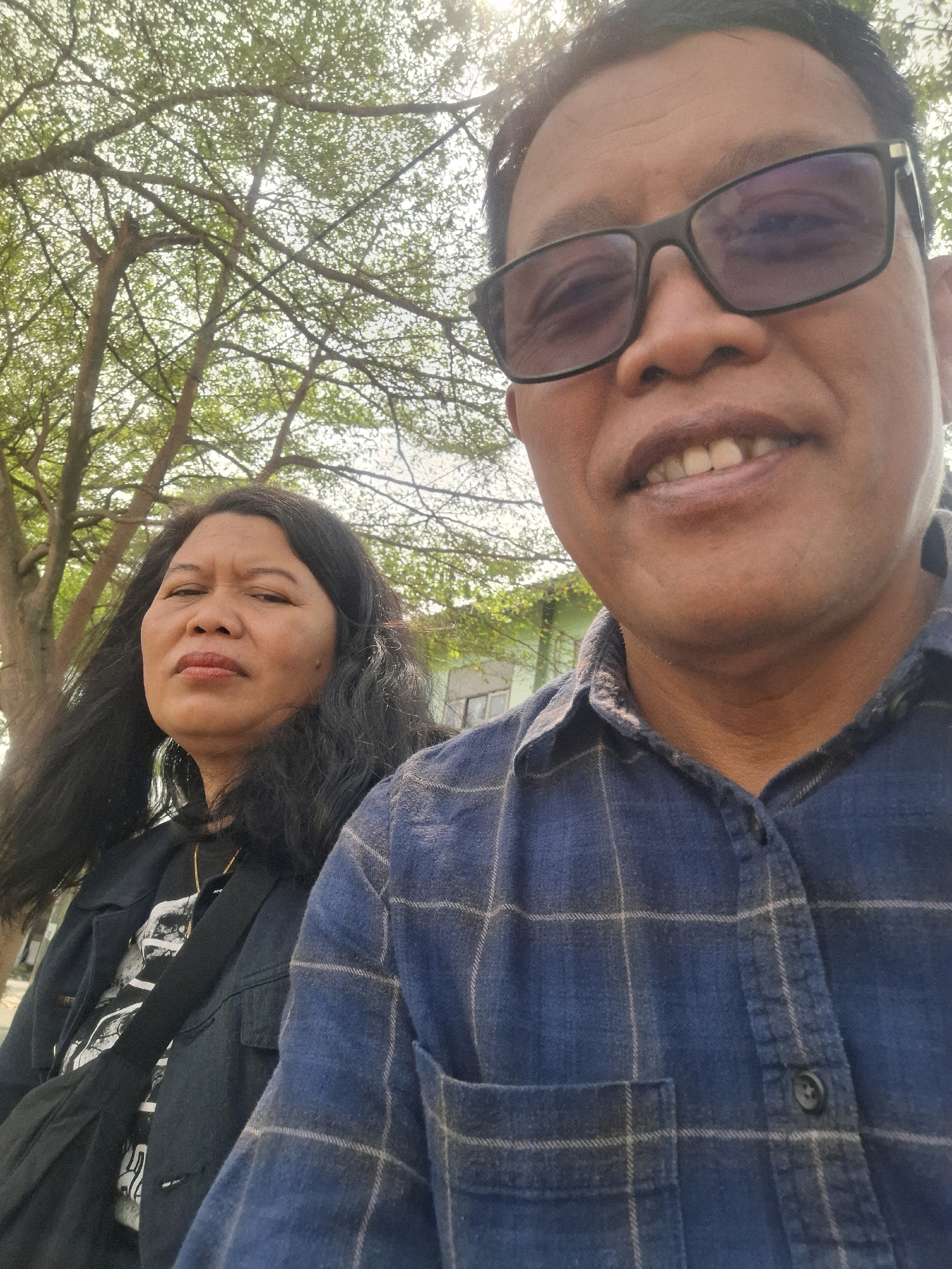 A selfie shows two adults, a man and a woman, in front of a building with trees in the background. The man, in the foreground, wears black-rimmed glasses and a blue plaid shirt. He is smiling, with his mouth open and teeth visible. The woman, positioned to the left of the man, has long black hair and a neutral expression. She is wearing a dark jacket over a dark shirt and a gold necklace. The scene appears to be outdoors, possibly on a street or in a park, with dappled sunlight filtering through the leaves of trees. The building in the background is light green.