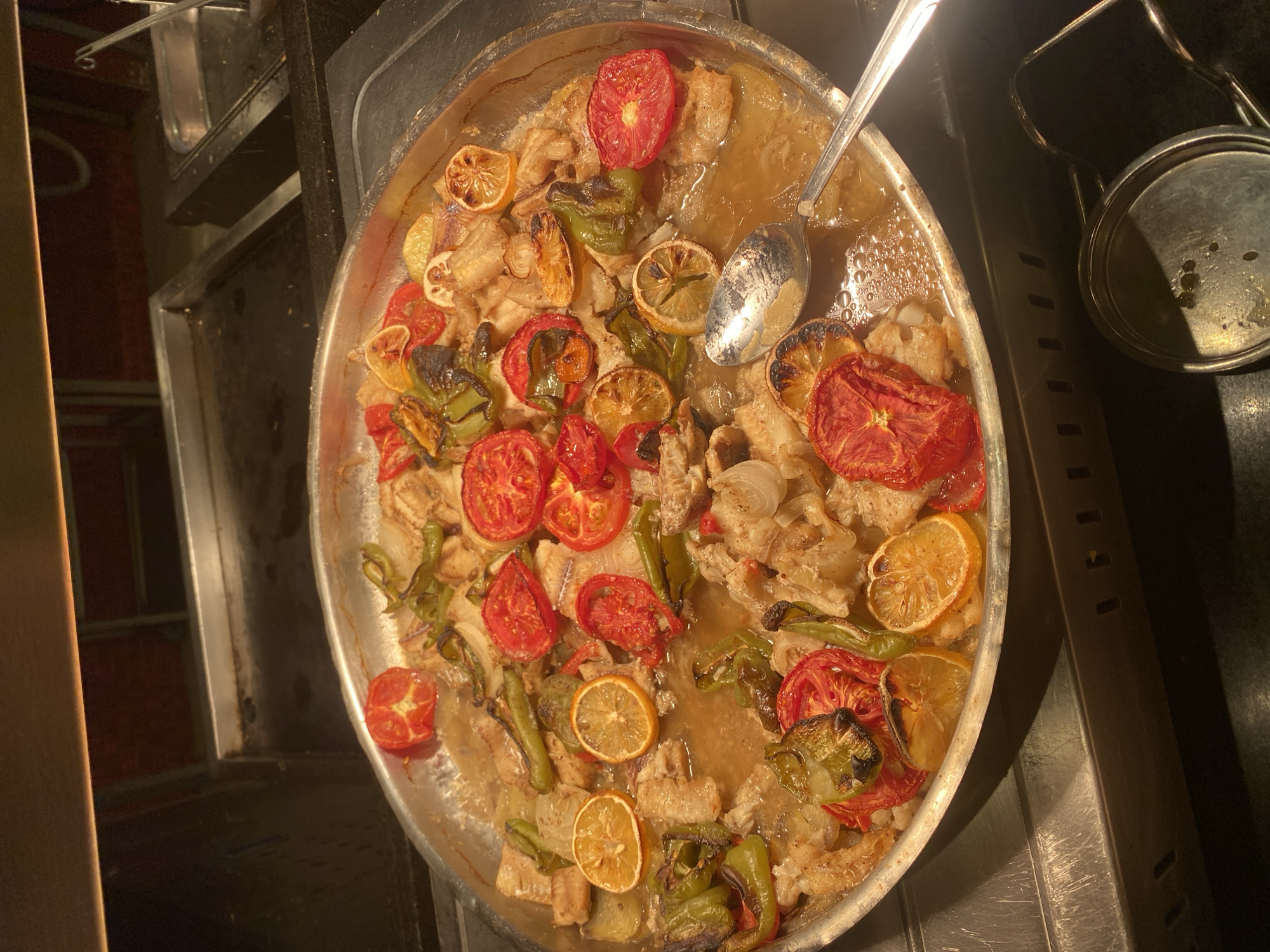 A large, round, metal pan is filled with a cooked fish dish, featuring chunks of white fish, sliced tomatoes, green peppers, onions, and lemon slices. The dish appears to be served from a buffet or a food service establishment. The pan is sitting on a stainless steel counter, which is part of a buffet setup. In the background, other stainless steel serving dishes and a warming element are visible. A silver spoon is resting in the pan, ready to serve the food. The lighting is warm and ambient, typical of a restaurant or hotel dining area. The scene is located in Aksu, Turkey.
