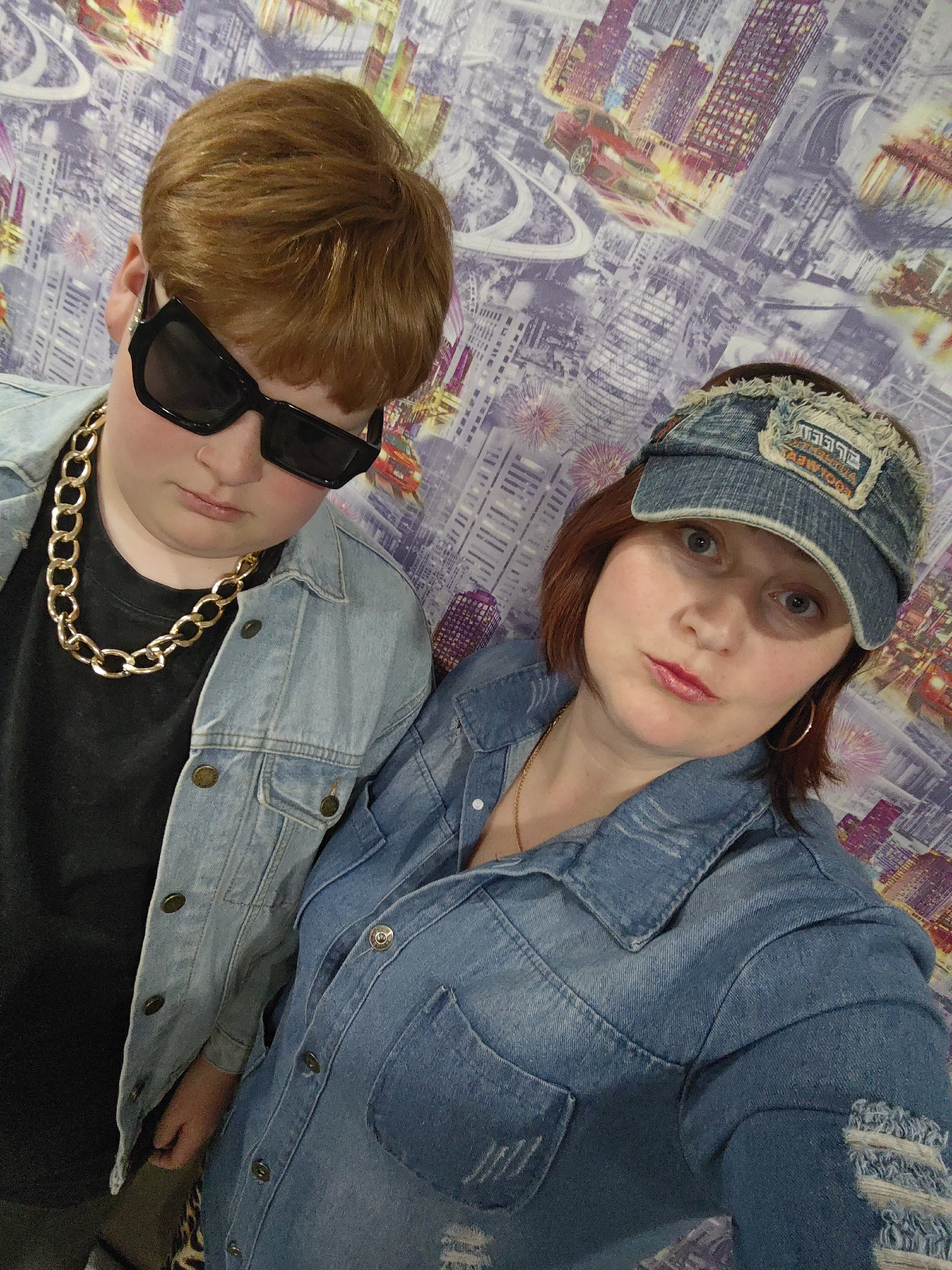 Two people pose for a selfie-style image. On the left, a person with short reddish-brown hair wears large black sunglasses, a gold chain necklace, and a denim jacket over a black shirt. On the right, a woman with reddish-brown hair wears a denim visor and a denim shirt-jacket. The background is a wall with a design resembling a cityscape with roads, buildings, and what appears to be a red car. The location is suggested to be in the city of Марковский, Russia.