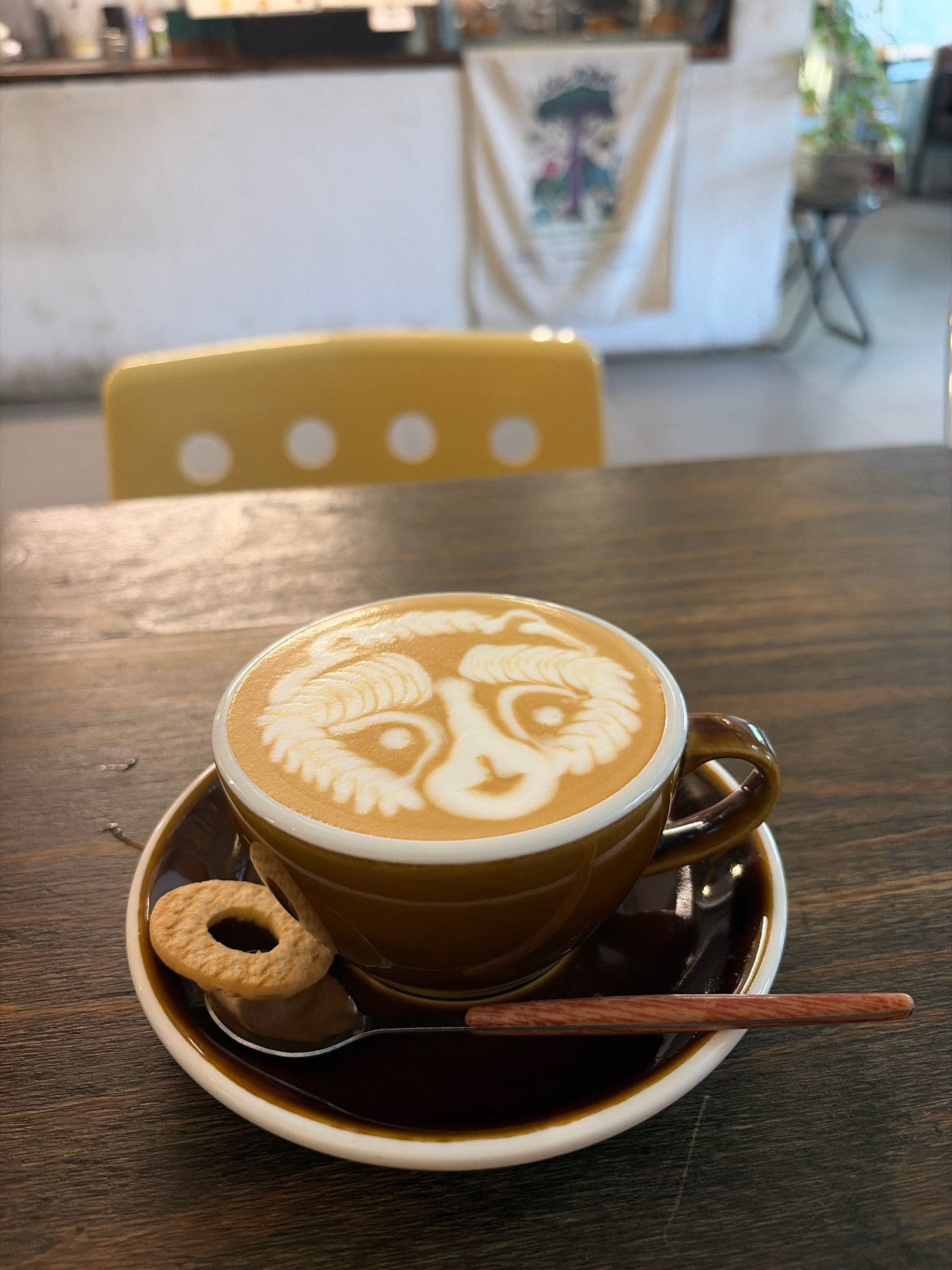 The image shows a close-up of a cup of coffee with latte art on a wooden table. The latte art depicts a face. A ring-shaped cookie and a wooden spoon are placed on the saucer. In the background, a yellow chair with white dots and a tapestry hanging on a wall are visible. The scene appears to be in a cafe in Đà Nẵng, Vietnam. There are no people visible in the image.