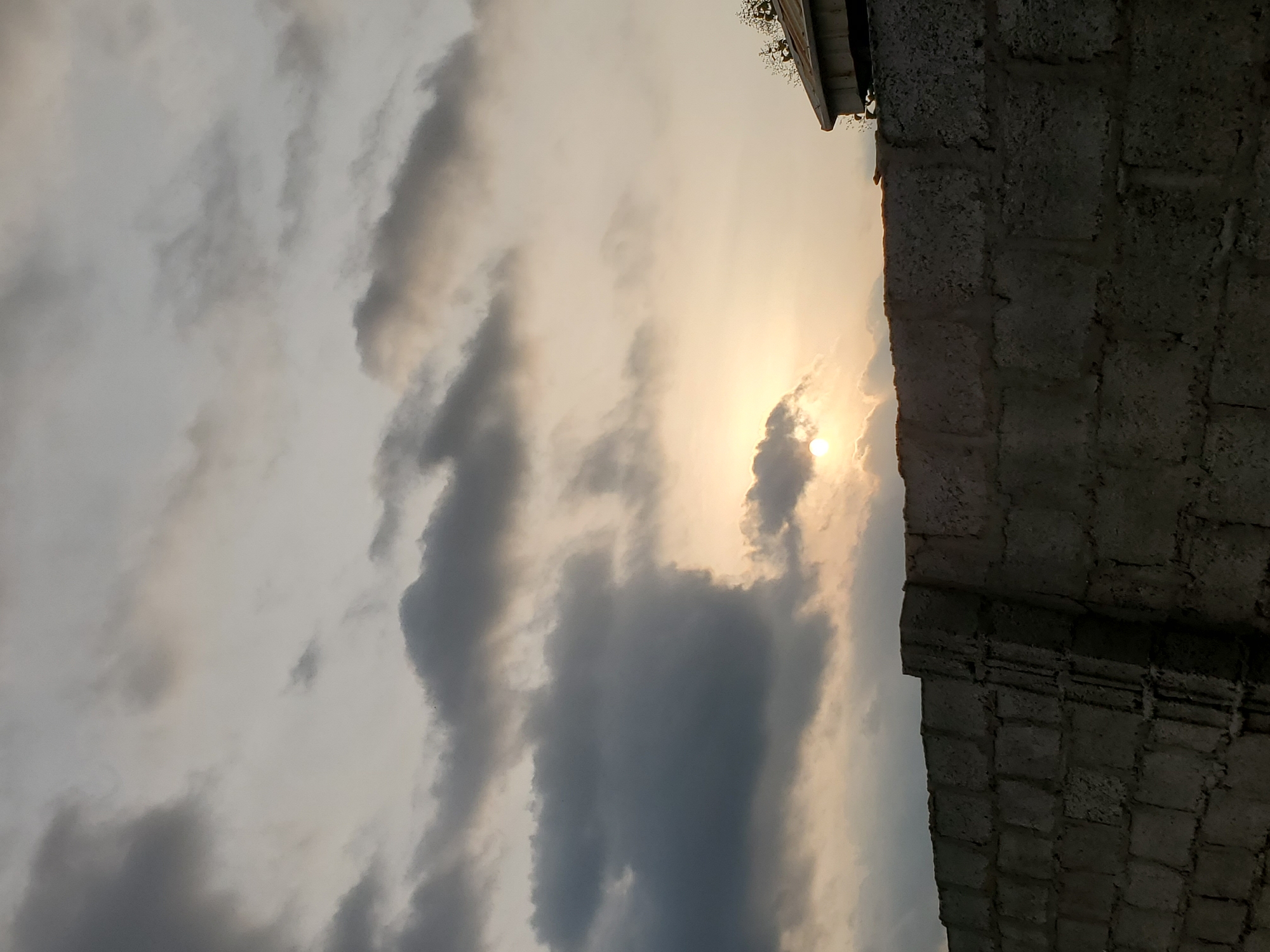 A low-angle, wide shot captures the sky over Suleja, Nigeria, as the sun sets. The sky is filled with a mix of dark, moody clouds and brighter, hazy patches where the sun is peeking through, casting a warm glow. A concrete block wall, constructed with grey blocks and visible mortar, runs horizontally across the lower portion of the frame, partially obscuring the view of the ground and any potential activity below. On the right side of the frame, a portion of a building with a slanted, metallic roof is visible, partially covered by foliage. The overall atmosphere is serene, with the natural light creating a dramatic backdrop for the man-made structures.