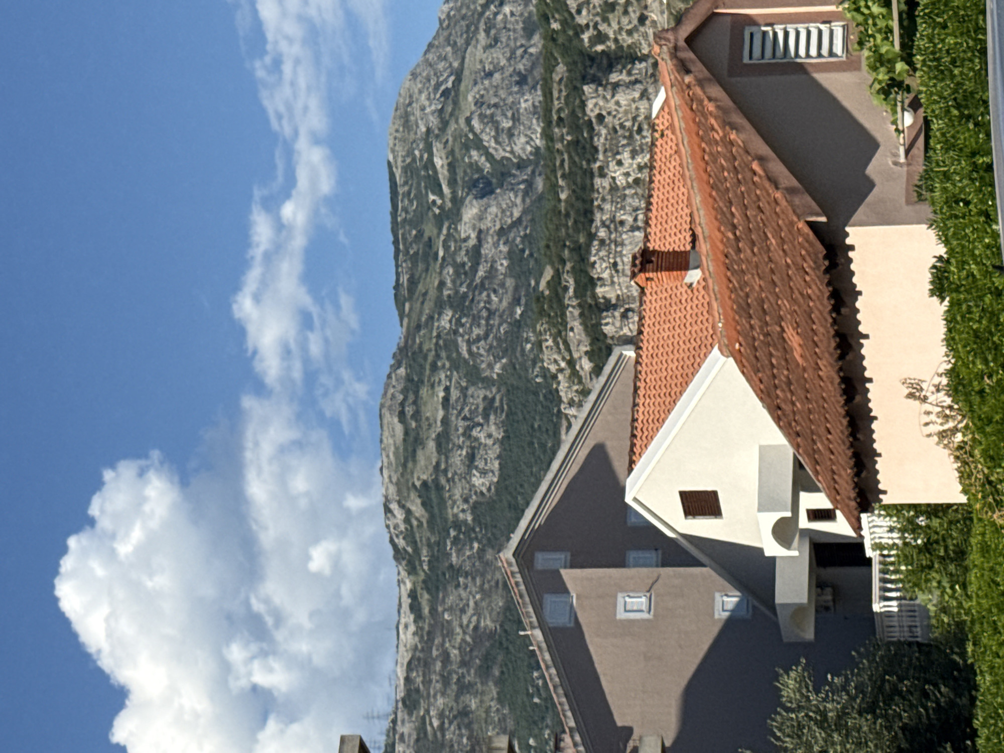The image is a photograph taken on a sunny day in Budva, Montenegro. It shows a view of residential buildings with red tiled roofs in the foreground, set against a backdrop of a rocky, sparsely vegetated mountain. The sky is blue with scattered white clouds. The buildings appear to be of a modern design with various architectural features, including balconies and windows. There are no people visible in the image. The overall impression is one of a Mediterranean coastal town with a mountainous landscape.