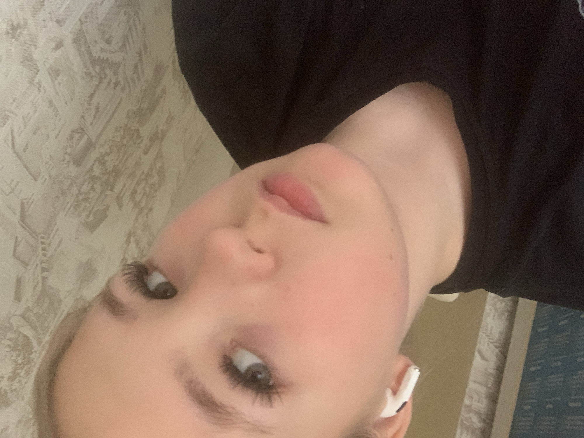 A close-up, slightly angled portrait shows a fair-skinned young woman with light blonde hair. She is wearing a dark-colored top, possibly a sweatshirt or hoodie. An earbud is visible in her right ear. Her facial features are prominent: light eyebrows, long eyelashes, and full lips. The background consists of a patterned wall, possibly wallpaper, and a glimpse of a surface with blue text or images on it. There is no information provided or visually evident within the image to suggest that this image was taken in the city of Chaikovsky, Russia.