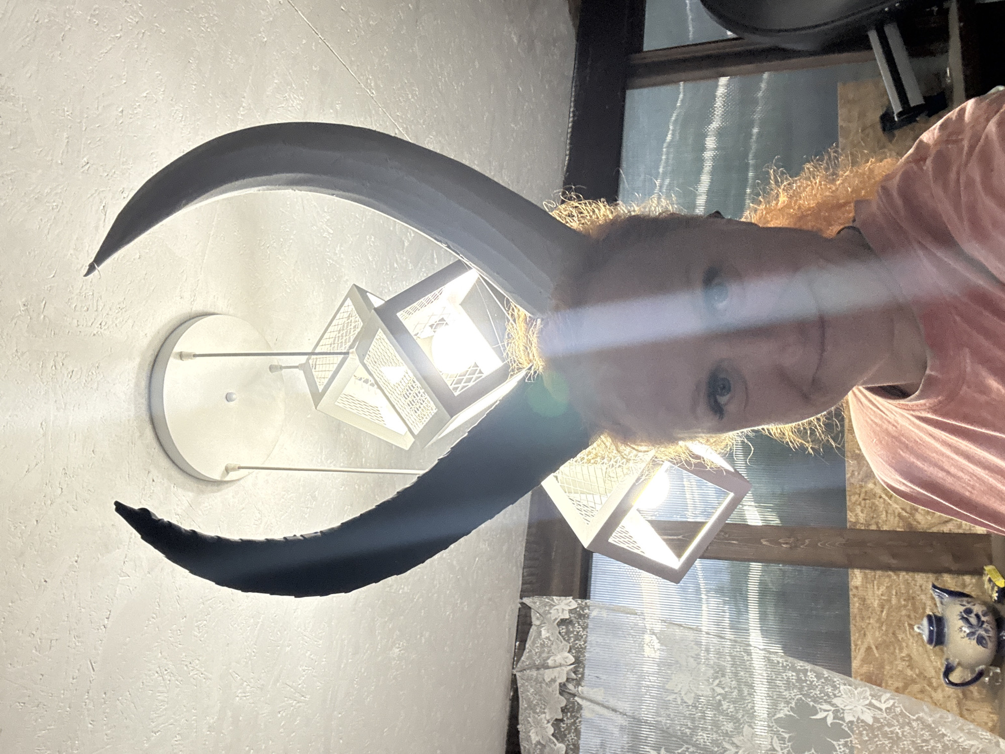 A woman with reddish hair is posing in front of a modern light fixture with two black curved horns, perhaps in a home in Chaikovsky, Russia. The light fixture has a circular base and three white cubic lamps. A window with a lace curtain and wooden frame is behind her, and a small blue and white teapot decoration is visible to the right.