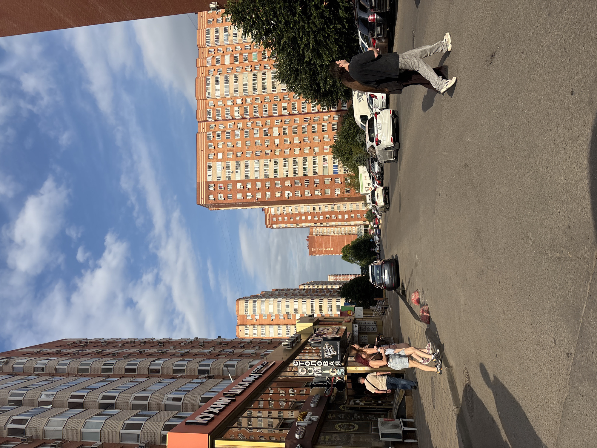 This image captures a street scene in Krasnodar, Russia, likely during daylight hours with a partly cloudy sky. On the left, a tall, modern apartment building with a light-colored brick facade and numerous balconies dominates the view. Its lower floors feature commercial storefronts with signage in Cyrillic, including "КУХНИ ШКАФЫ" and "СТОЛОВАЯ." Several people are visible on the sidewalk in front of these shops, including a man walking away from the camera and two women standing near the entrance.
Across the street, a wide asphalt road stretches into the distance, lined with parked cars on both sides. Another row of identical, tall, reddish-orange apartment buildings can be seen on the right side of the road, also featuring many balconies. A white van is also parked in the street. Further down the road, more apartment buildings are visible, receding in perspective.
In the foreground, a couple is crossing the street, with one person in a black t-shirt and grey pants walking ahead of another person. The overall impression is of a typical urban residential and commercial area in Russia.