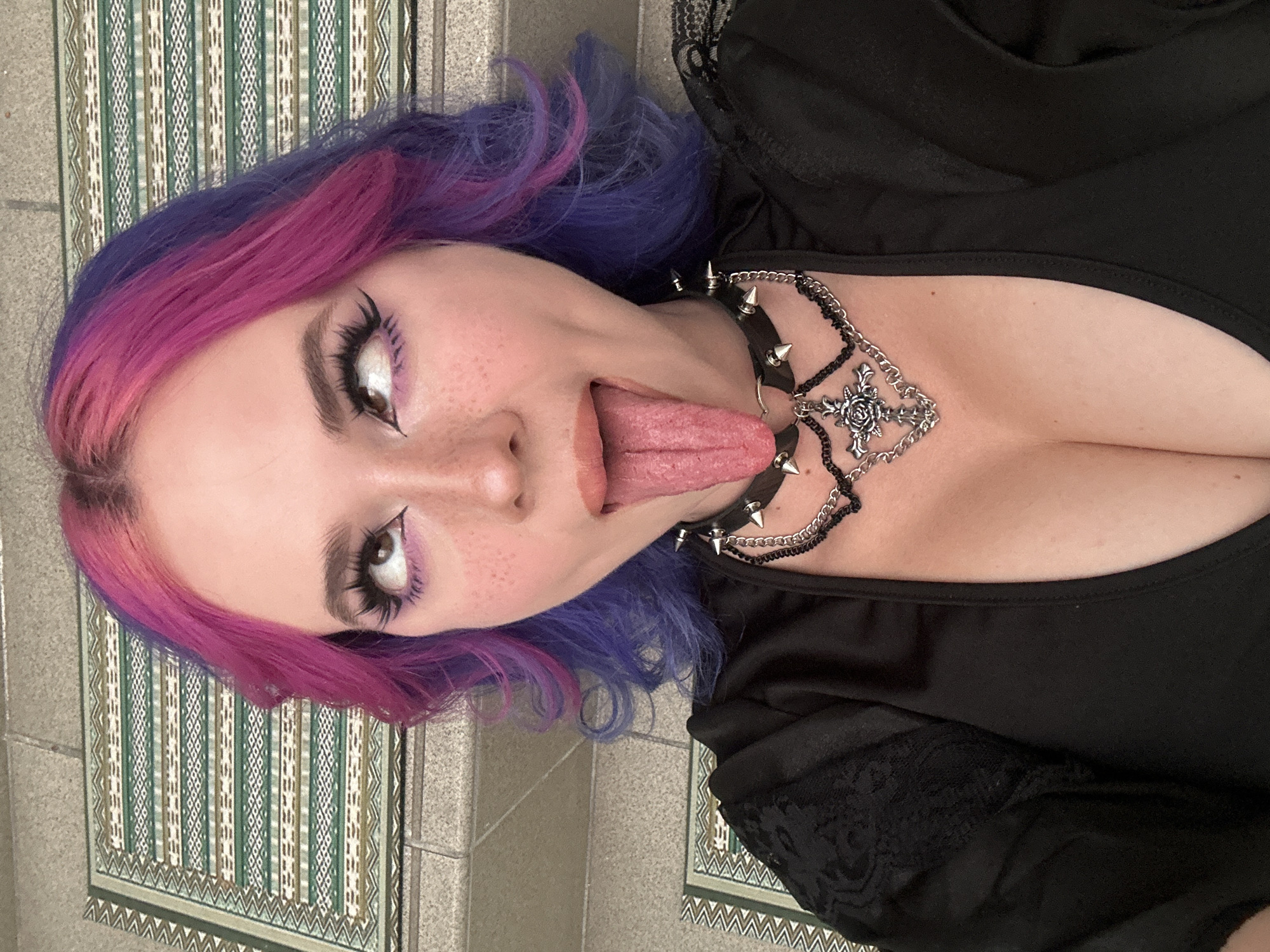 A close-up, rotated shot depicts a person with striking two-toned hair, vibrant pink blending into purple. The individual has a fair complexion with visible freckles and is playfully sticking out their tongue. Their eyes are accentuated with dramatic winged eyeliner and purple eyeshadow. They are wearing a black top with delicate lace trim visible on the shoulder. Around their neck, they sport a spiked black choker and a layered silver necklace featuring a prominent gothic pendant, possibly a rose within a cross. The background consists of patterned floor tiles with green, white, and brown geometric motifs, set against plain grey-beige tiled walls, suggesting an indoor setting in the City of Zagreb, Croatia. No text is visible in the image. The overall impression is one of expressive, alternative style.
