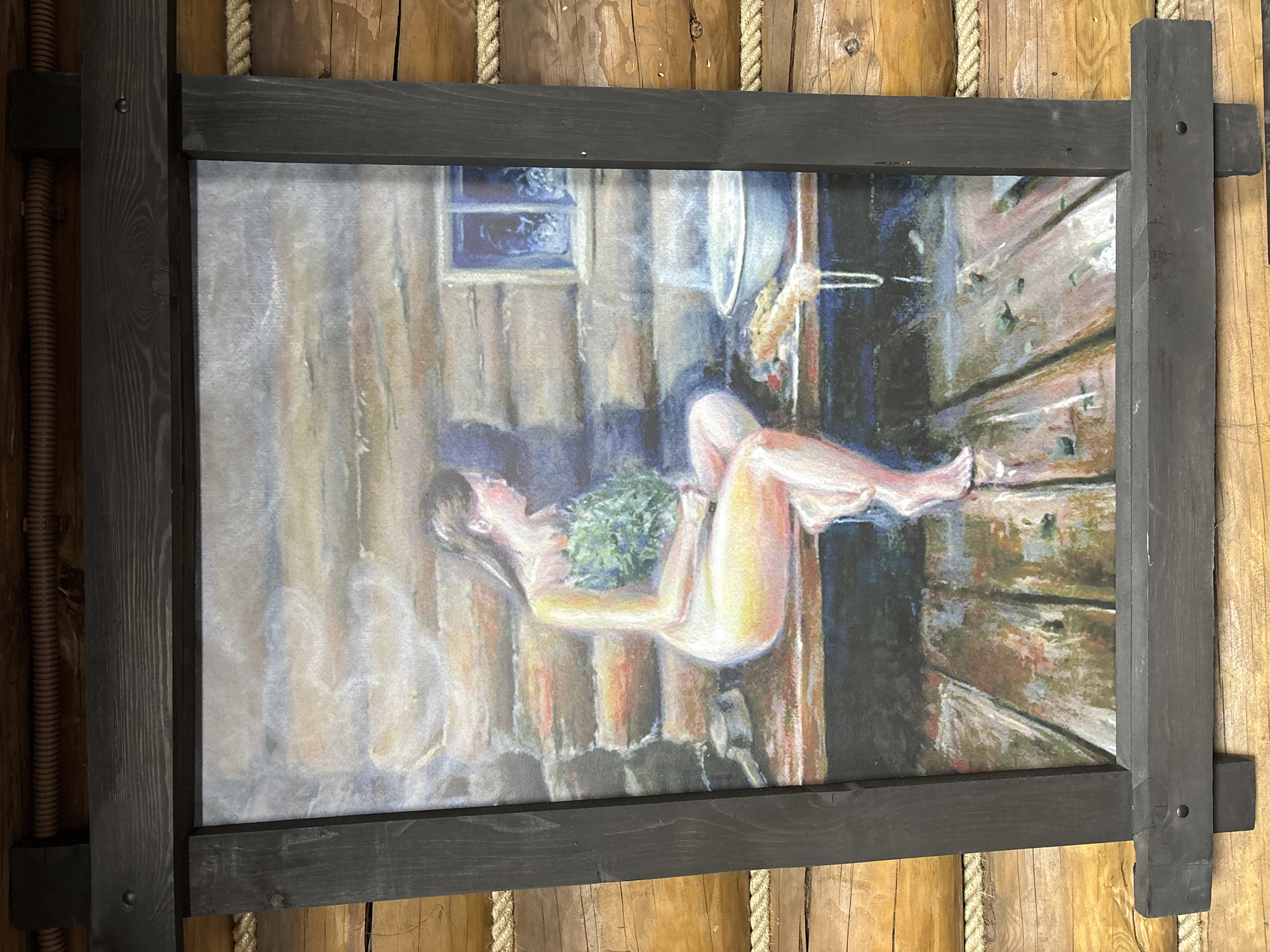 An image of an impressionist style painting framed in black wood, hung on a wall of rough, unpainted wooden planks and beams. The painting depicts a nude woman lying down in what appears to be a sauna or similar wooden structure. She is holding a bundle of green leafy branches or herbs. There is a window visible, and a metal basin or bucket hangs from a wall. The scene evokes a sense of warmth and steam. The overall location seems to be in Prikamskii, Russia, as suggested by the style and content of the painting, which is often associated with Russian bathing traditions.