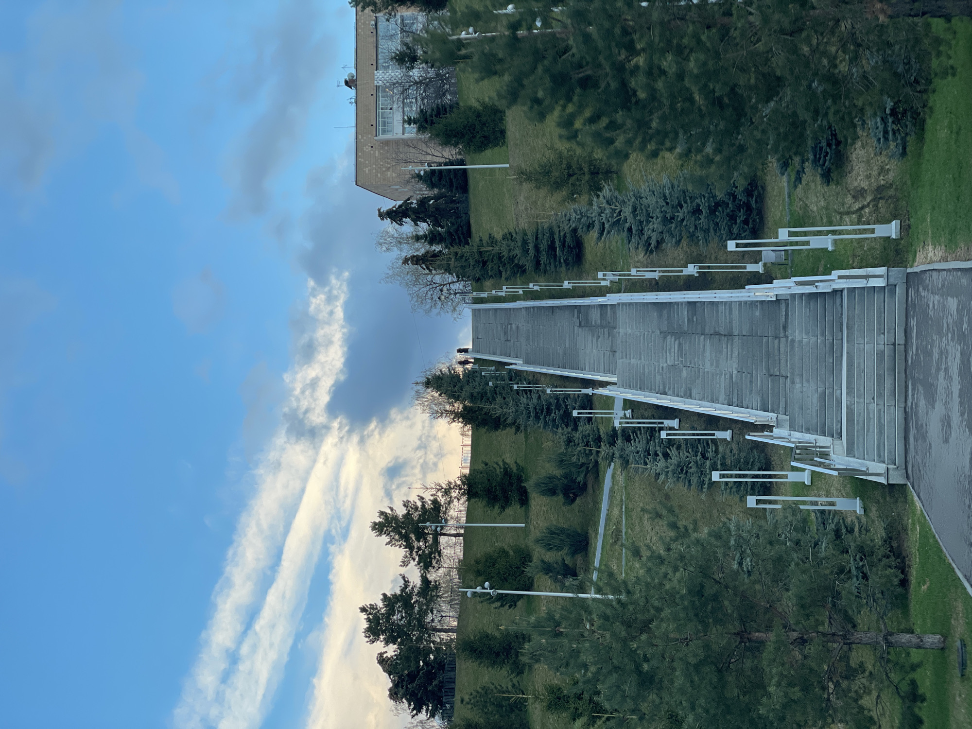The image captures an outdoor, multi-level urban park or landscaped area under a partly cloudy sky. The perspective is slightly elevated, looking across the scene.
A prominent feature is a wide, grey staircase with multiple landings, ascending diagonally from the lower-left to the mid-right of the frame. Modern, rectangular white light fixtures or railings line both sides of this staircase. To the right of the staircase, a paved pathway runs parallel, suggesting an alternative route.
The ground comprises manicured grassy slopes and lawns, densely planted with various trees and shrubs. There are numerous tall evergreens, including pines and spruces, providing a rich green and bluish-green palette. Some deciduous trees, appearing bare, are also visible, suggesting the season might be late autumn, winter, or early spring. Several white streetlights, matching the style of the staircase fixtures, are positioned throughout the grassy areas.
In the upper-left background, a multi-story building with a light-colored facade and visible windows is partially seen, hinting at an urban or institutional setting. Antennae or satellite dishes are visible on its roof.
The sky occupies a significant portion of the upper frame, displaying a clear blue hue with scattered clouds. Some clouds are thin and elongated, stretching across the blue expanse, while others are thicker and darker grey. The lighting suggests it is daytime, possibly morning or late afternoon given the quality of light and cloud formations.
There are no people or discernible activities taking place. No visible text is present in the image.