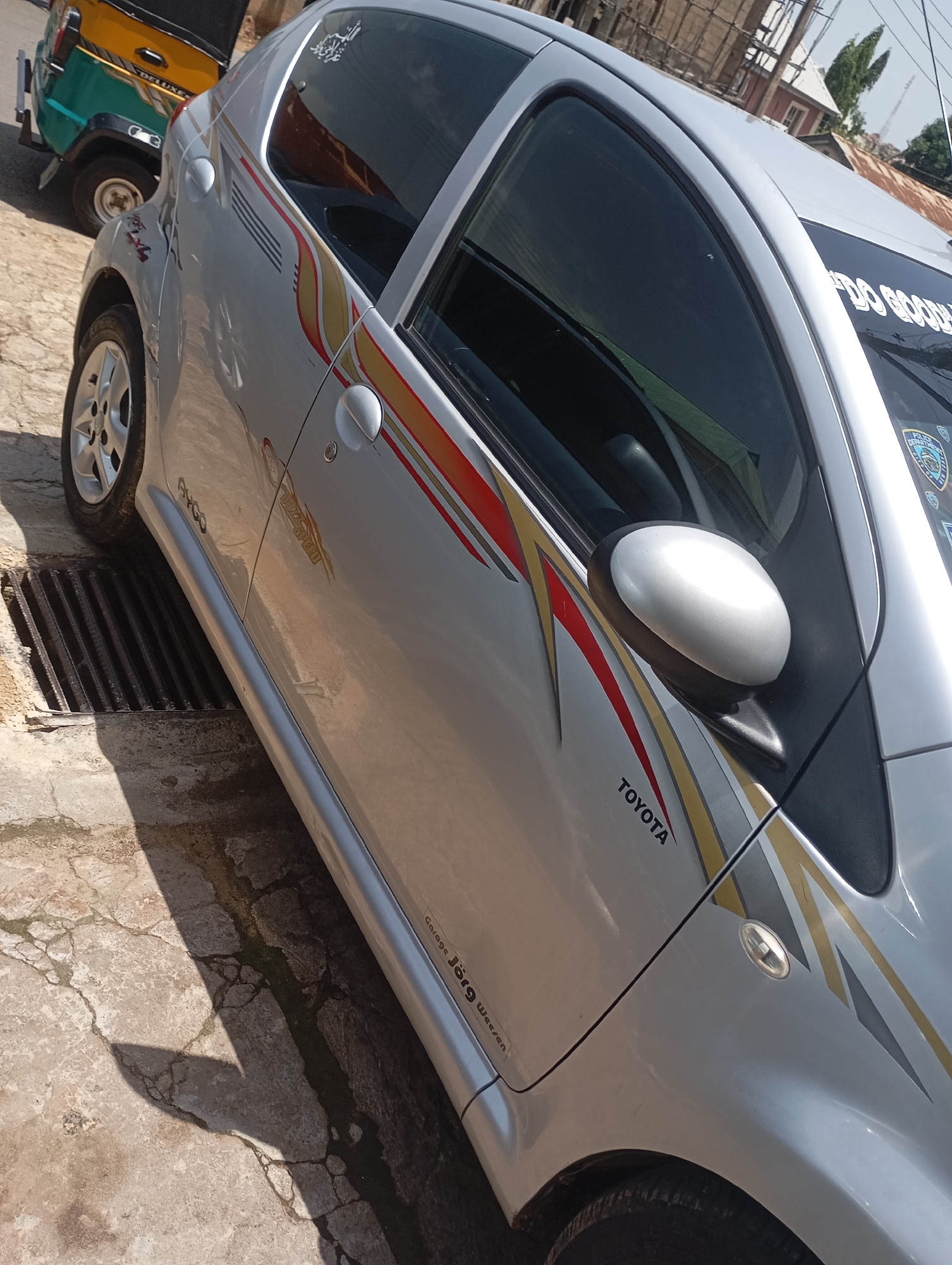 A silver Toyota Aygo is parked on a cracked concrete surface next to a metal grate. The car features decorative decals with red, gold, and gray stripes on its side. The car's windows are tinted. A yellow and green three-wheeled auto-rickshaw is visible in the background, parked to the left of the car. The scene appears to be an outdoor setting, possibly a street or parking area. The lighting suggests it is daytime, with a shadow of an unseen object falling on the ground. Text on the car includes "TOYOTA" and "Garage Jörg Wesen". There is also text on the windshield, partially obscured, that reads "DO GOOD". Additionally, a police department sticker is visible on the windshield. The location context is Jos, Nigeria.