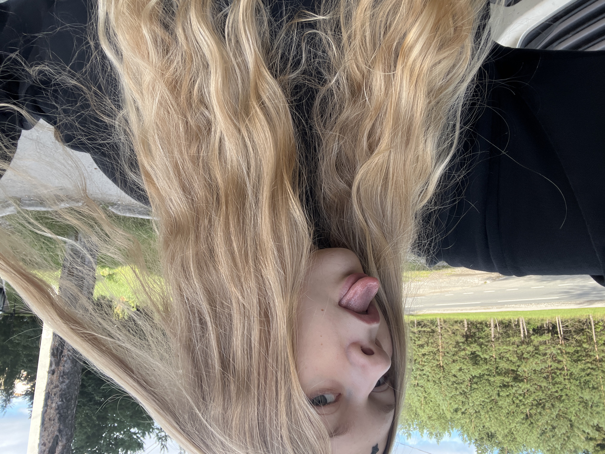 A young person with long, blonde hair is shown from the chest up, outdoors. They are sticking out their tongue and holding a strand of their hair to their right. A black garment is worn. In the background, a road is visible, bordered by a line of evergreen trees. The sky is cloudy. The image appears to have been taken in Perm, Russia, given the context.