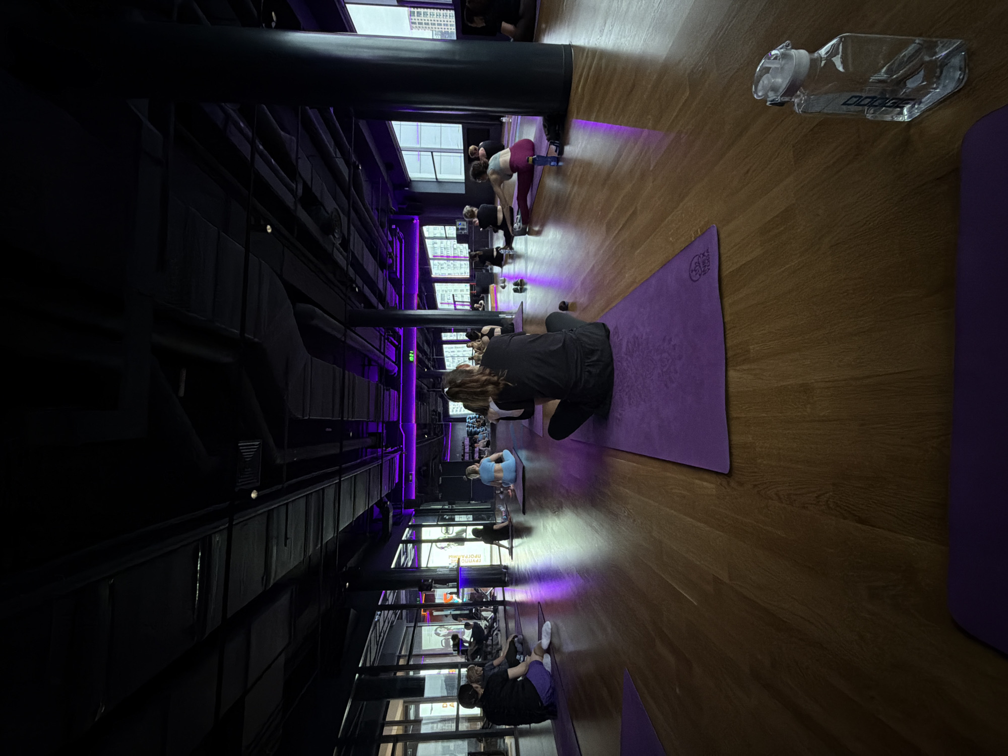 The image shows an interior scene, possibly a yoga or fitness studio. A group of people are positioned on yoga mats along the length of the room. The people are engaged in various stretches or exercises, indicating a class in progress. The studio has a modern, industrial design with exposed black metal beams and ductwork in the ceiling. Tall windows line one side of the room, offering a view of buildings outside. The lighting is a combination of natural light and purple-toned artificial lighting. There is a water bottle on the wooden floor. The location is possibly in Moscow, Russia, based on the architecture of the visible buildings outside the windows.