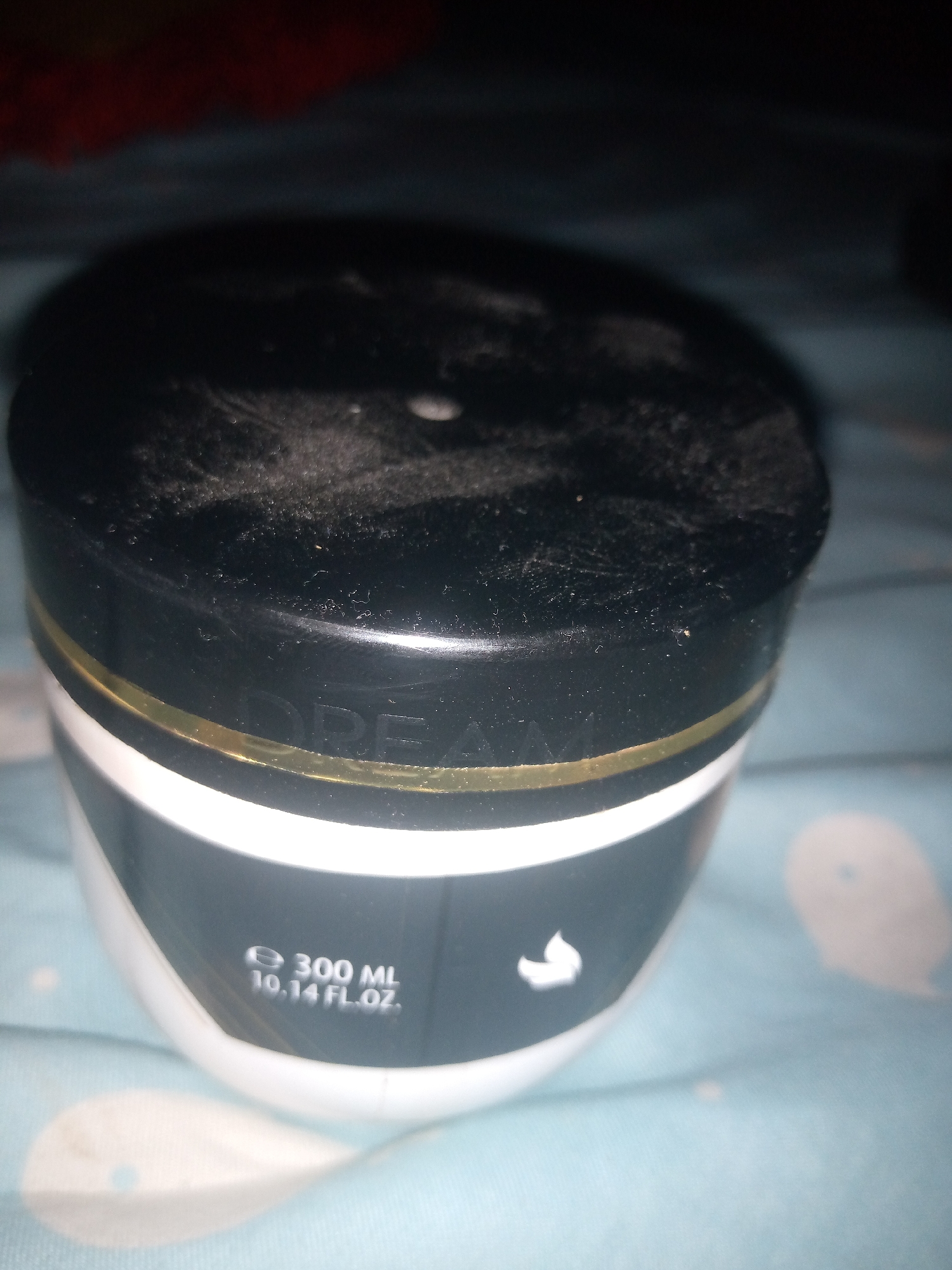 A jar with a black lid is placed on a patterned surface. The jar has a gold band around its neck, and white text on the body reads "e 300 ML 10.14 FL.OZ.". The lid appears dusty. The background is a blue and white patterned cloth. There is a red object in the upper left corner, out of focus.