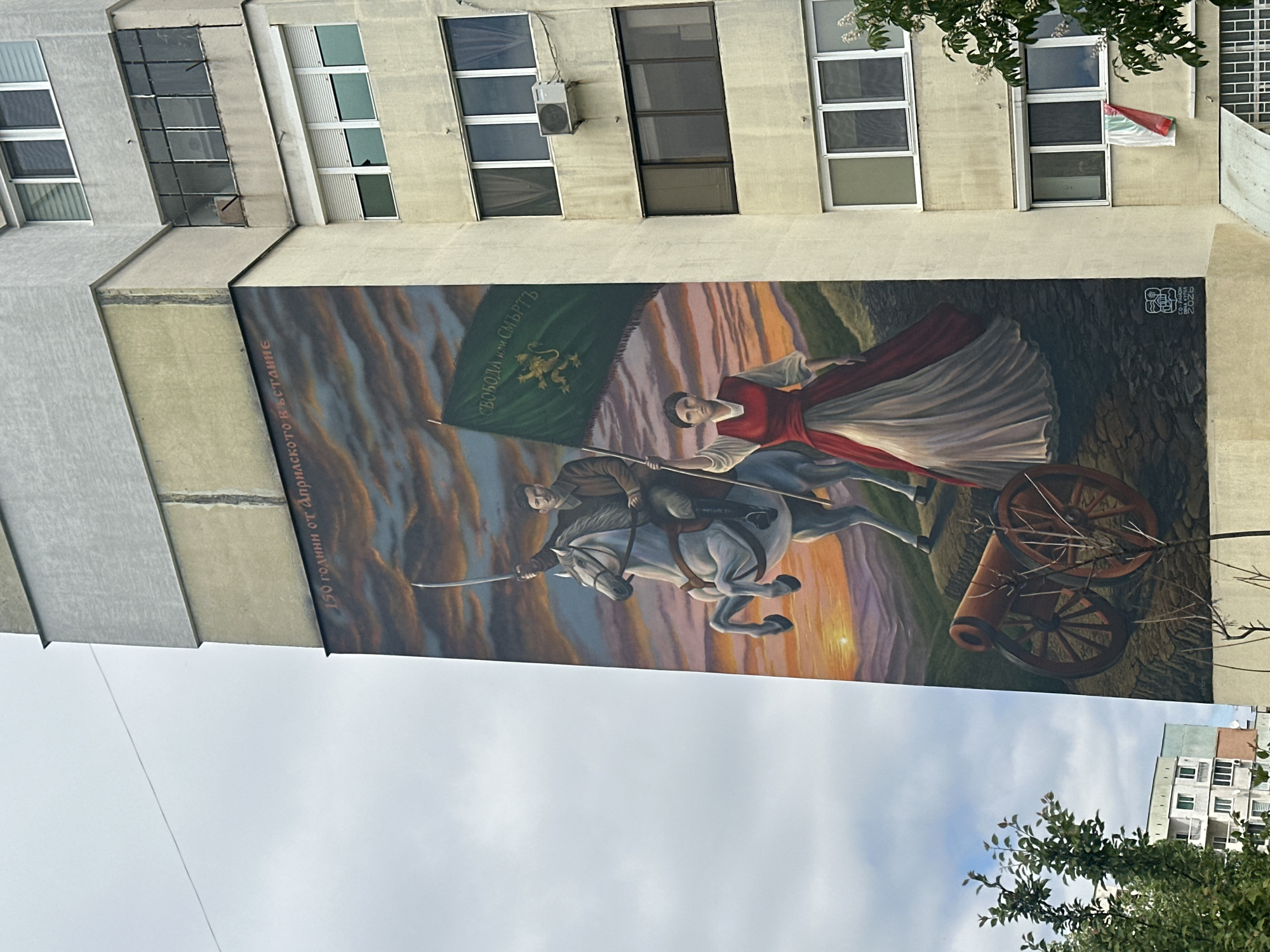 A mural depicts a man on horseback carrying a flag, with a woman beside him in a red and white dress. A cannon is positioned to the right. The mural is painted on the side of a multi-story building, with several windows visible above it. The flag features a lion and the text "СВОБОДА или СМЪРТЬ." The text "150 години от Априлското въстание" is also present. The mural is located in an unknown city.