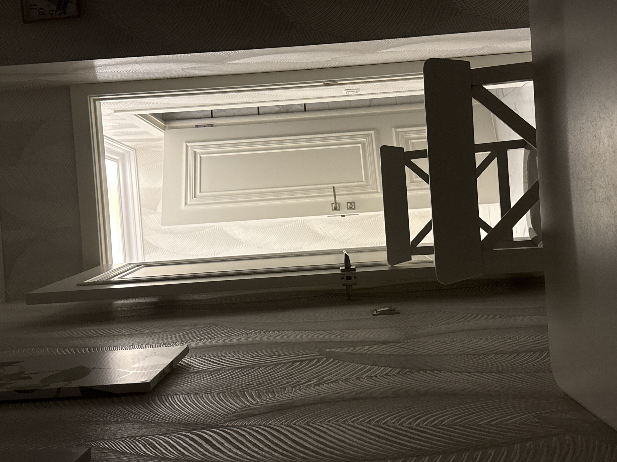 The image appears to be an interior shot, possibly a hallway or room with a doorway leading to another space. The perspective is unusual, as if the camera is positioned very low to the ground and looking upwards.
In the foreground, there is a textured surface, likely a wall or floor, with a repeating pattern that resembles waves or leaves. A flat, rectangular object with a patterned surface is placed on this textured surface, angled towards the viewer.
The central focus of the image is a doorway with a white-framed door. The door itself is open, revealing a glimpse of another room beyond. This second room also features white-trimmed elements, suggesting a consistent design theme. A light source, possibly a window, is visible in the second room, illuminating it with a bright glow. A part of a wooden structure, possibly a gate or railing, is visible to the right of the doorway, casting a shadow.
At the top of the frame, a portion of a clock face is visible, showing the numbers 10, 11, and 6. The hands of the clock are not clearly discernible. The time appears to be sometime between 10 and 11 o'clock, but the exact hour and minute are uncertain due to the angle and partial view. The lighting suggests it could be daytime, with artificial light also contributing to the illumination. The overall atmosphere is somewhat dimly lit in the foreground, contrasting with the brighter space visible through the doorway.
There is no visible text beyond the numbers on the clock face. The scene does not contain any people or animals. The location context provided, Derbent, Russia, is not visually evident from the image itself.