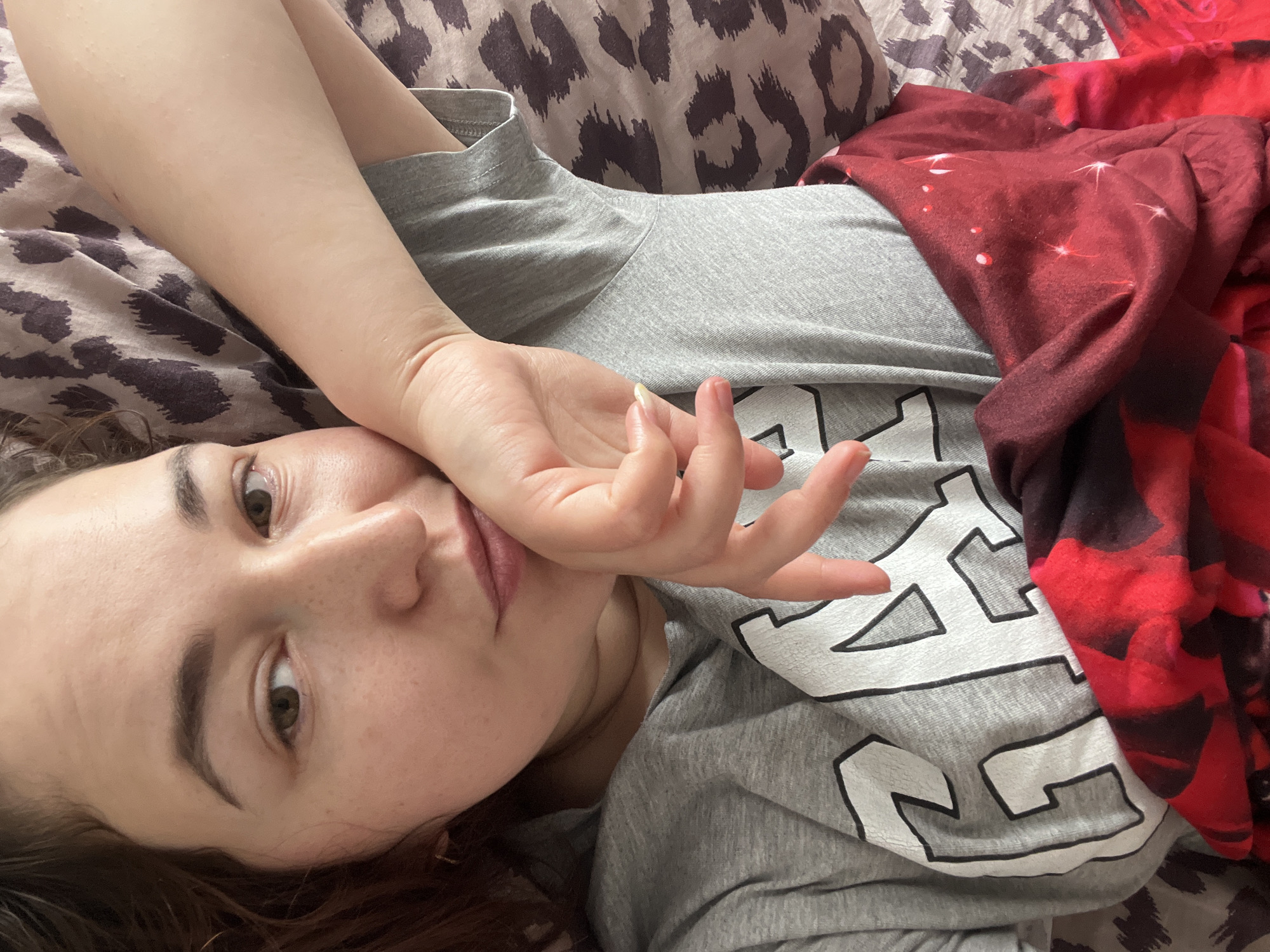 An overhead medium shot shows a young woman lying on her back on a bed. The bed is covered with patterned sheets, and a red blanket with sparkling patterns is draped over her legs. The woman is wearing a grey t-shirt with large white letters on it. Her right arm is bent at the elbow, and her hand is resting on her forehead. Her head is tilted to the left, and she is looking at the camera. Her eyes are open, and her lips are slightly parted. The overall impression is one of relaxation and comfort. The image does not contain any information that would allow for its localization in Chelyabinsk, Russia.