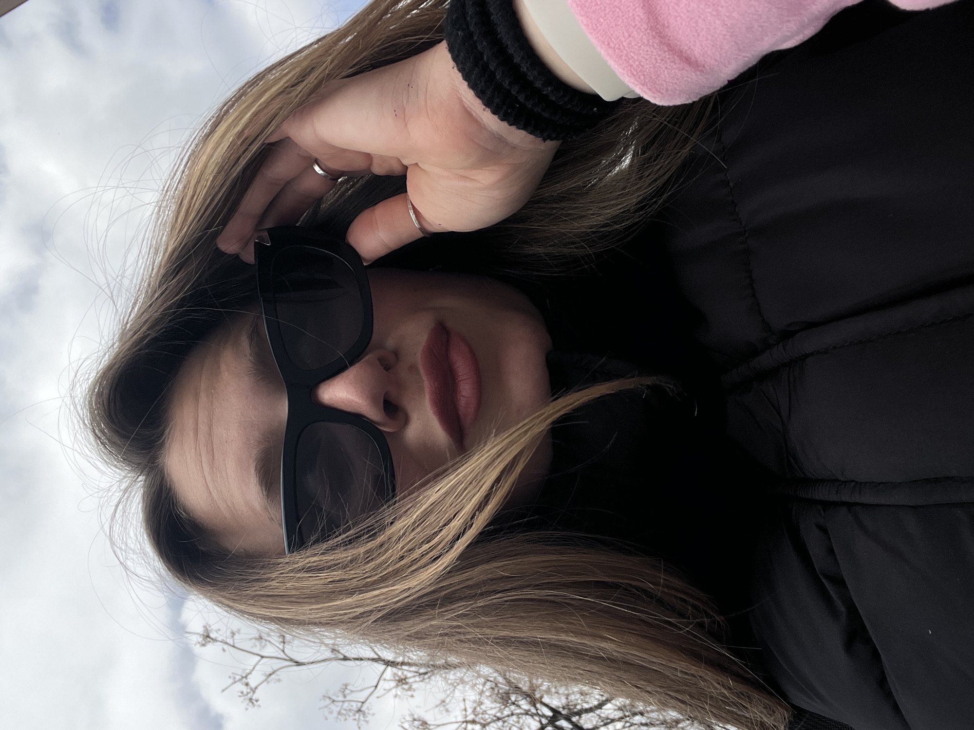 A selfie shows a fair-skinned person with long brown hair and wearing dark sunglasses. The person is wearing a pink and white sleeved top with a black woven bracelet on their wrist and a ring on their finger, and a black puffer jacket. The background shows a cloudy sky and bare tree branches. It is not possible to identify the location in Budva, Montenegro from this image alone.