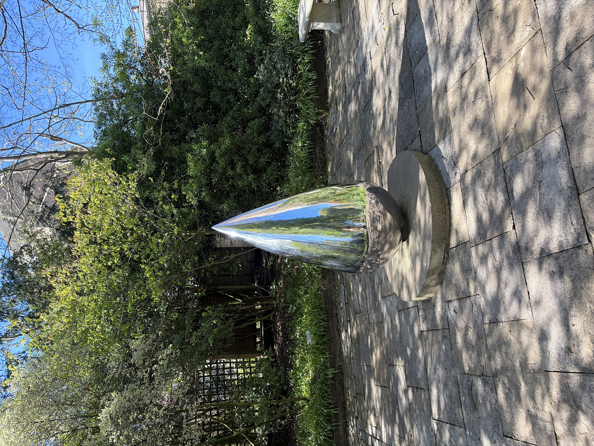 The image shows a shiny metal sculpture in a garden or park. The sculpture appears to be conical, resting on a circular stone base. The sculpture reflects the surrounding environment, including trees and sky. The ground is paved with rectangular stones, casting shadows. The location is West Suffolk, United Kingdom.