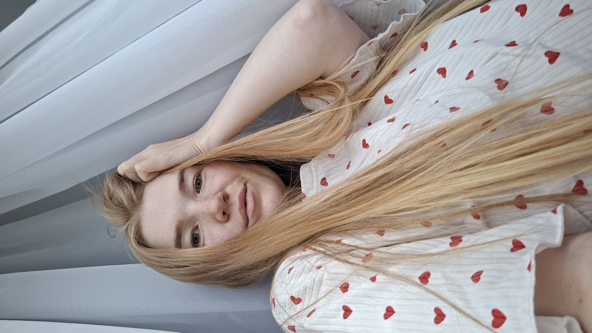 A young, fair-skinned woman with long, light blonde hair lies on what appears to be a bed or soft surface. She is wearing a white top or dress with small red heart patterns. One hand is resting near her head, possibly touching her hair. The background includes white, translucent fabric, suggesting a window curtain or draped material. The location (Chaikovsky, Russia) cannot be determined from the visual information in the image.