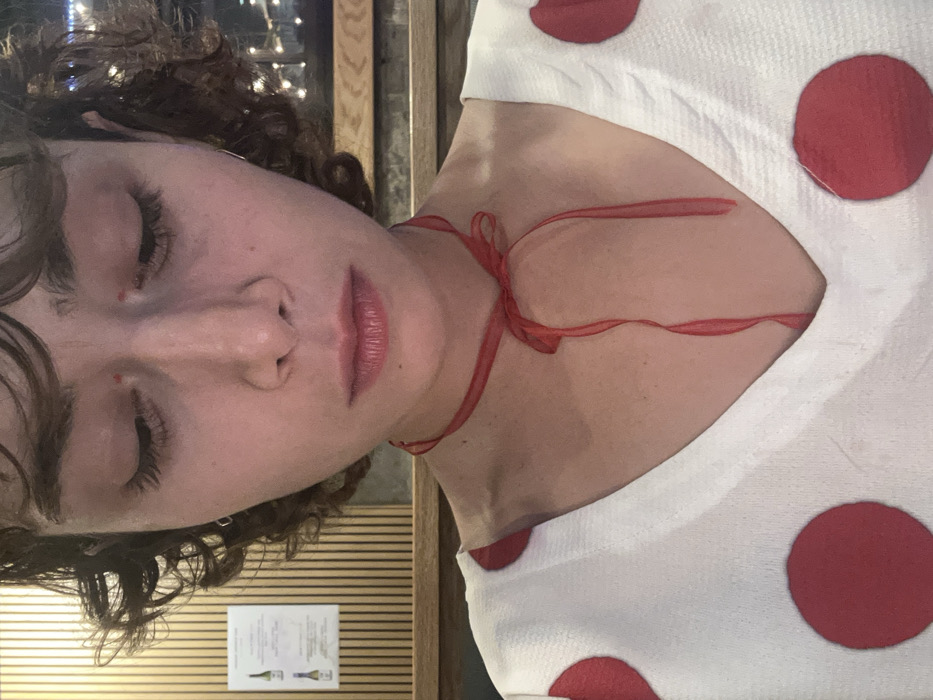 A person with curly brown hair and long eyelashes is tilted to the side, their eyes closed. They are wearing a white top with large red polka dots. A thin red ribbon is tied loosely around their neck, with the ends draped across their chest. A beige wall with a vertical wood design and a small sign is visible in the background on the left. Part of a window with blurred lights appears in the top center.