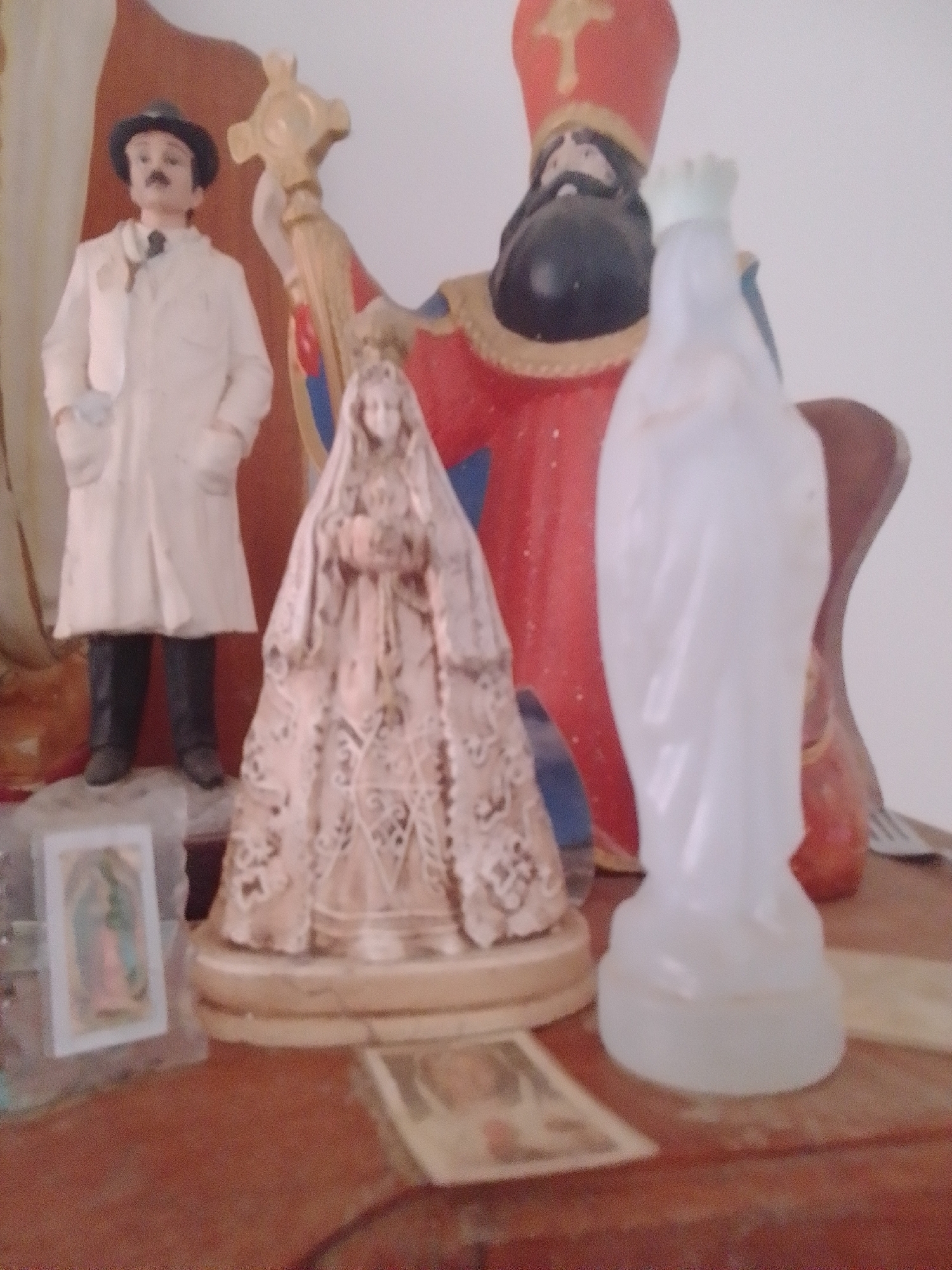 This media file is a close-up, low-angle shot of a collection of religious figurines displayed on a wooden surface, likely a shelf or altar. In the background, a larger figurine of a person in a red robe and a tall, red miter hat stands prominently. To the left of this, a figurine of a man in a white coat, black hat, and black shoes stands with his hands in his coat pockets. Between these two larger figures, a detailed, light-colored figurine of a robed woman holding an object to her chest is positioned. In front of the central figures, a white, opaque figurine of a robed figure, possibly another religious statue, stands with its hands extended forward. To the far left, a small, rectangular holy card depicting the Virgin Mary is visible, partially obscured by another object. The overall scene is one of a devotional display, possibly in a home or a small chapel in La Tagua, Venezuela. The lighting is soft, suggesting an indoor setting during daylight hours. The figurines are the primary subjects, and their arrangement suggests a devotional or decorative purpose. There is no visible text in the image.