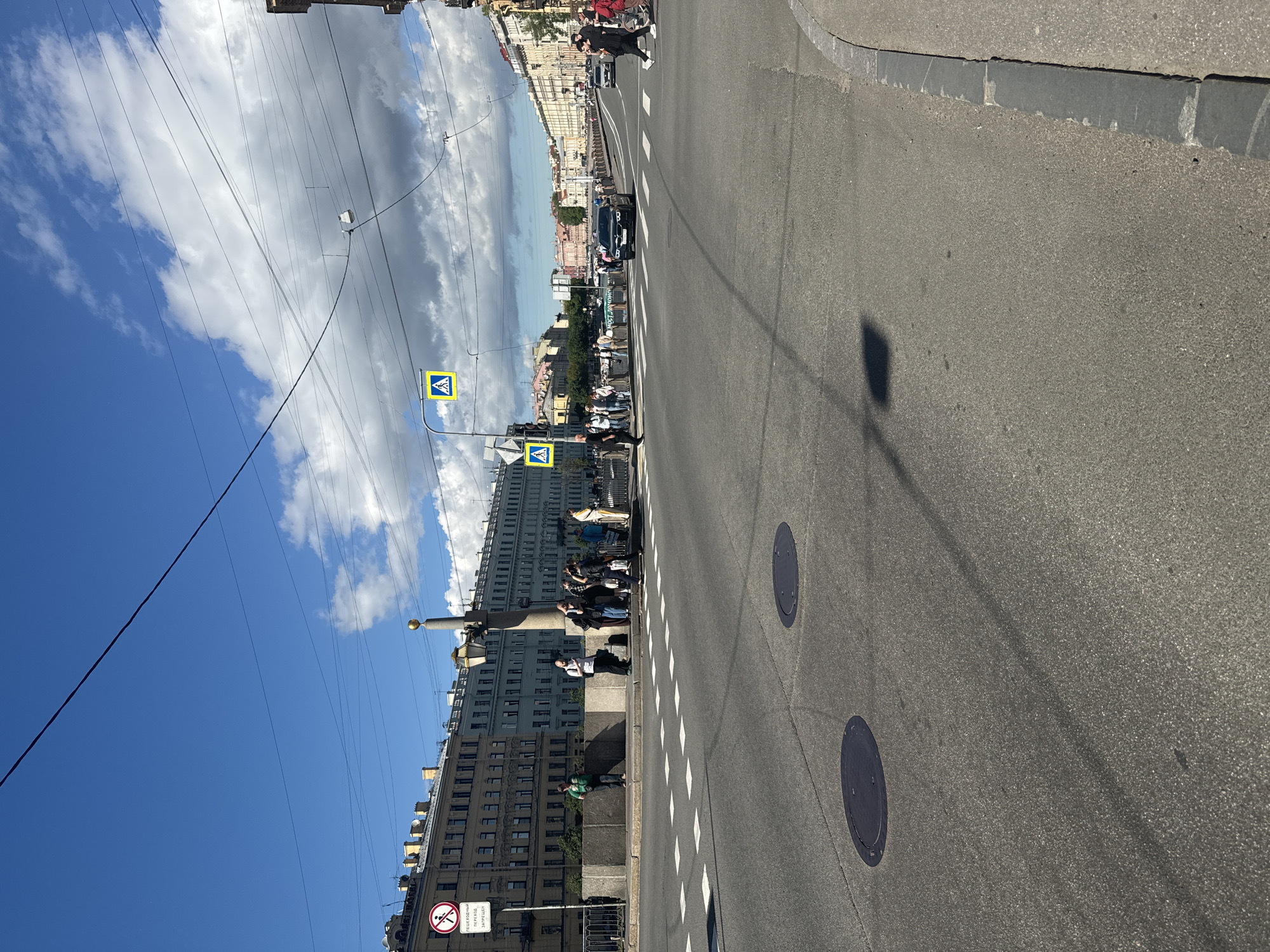The image captures a street scene in Saint Petersburg, Russia, likely on a bridge due to the presence of a canal or river in the background. The perspective is from the street level, looking across an intersection. The sky is blue with scattered clouds, and power lines are visible overhead.
On the left side of the image, there is a multi-story building with a classical architectural style, typical of Saint Petersburg. A pedestrian crossing is marked on the road with white dashed lines. Several pedestrians are visible on the sidewalk and near the crossing, waiting to cross or walking. A sign with a red circle and a diagonal line through a pedestrian symbol, indicating "no pedestrian crossing," is mounted on a pole near the building.
In the center of the image, a black car is driving through the intersection. There are more buildings and a bridge with railings visible in the distance, lining the waterway. Pedestrian crossing signs with yellow backgrounds and white pedestrian symbols are present at the intersection.
On the right side, the road curves and continues into the distance. Another car is visible further down the road. The street has concrete curbs and manhole covers are present on the asphalt.
The overall impression is a lively urban environment on a clear day.