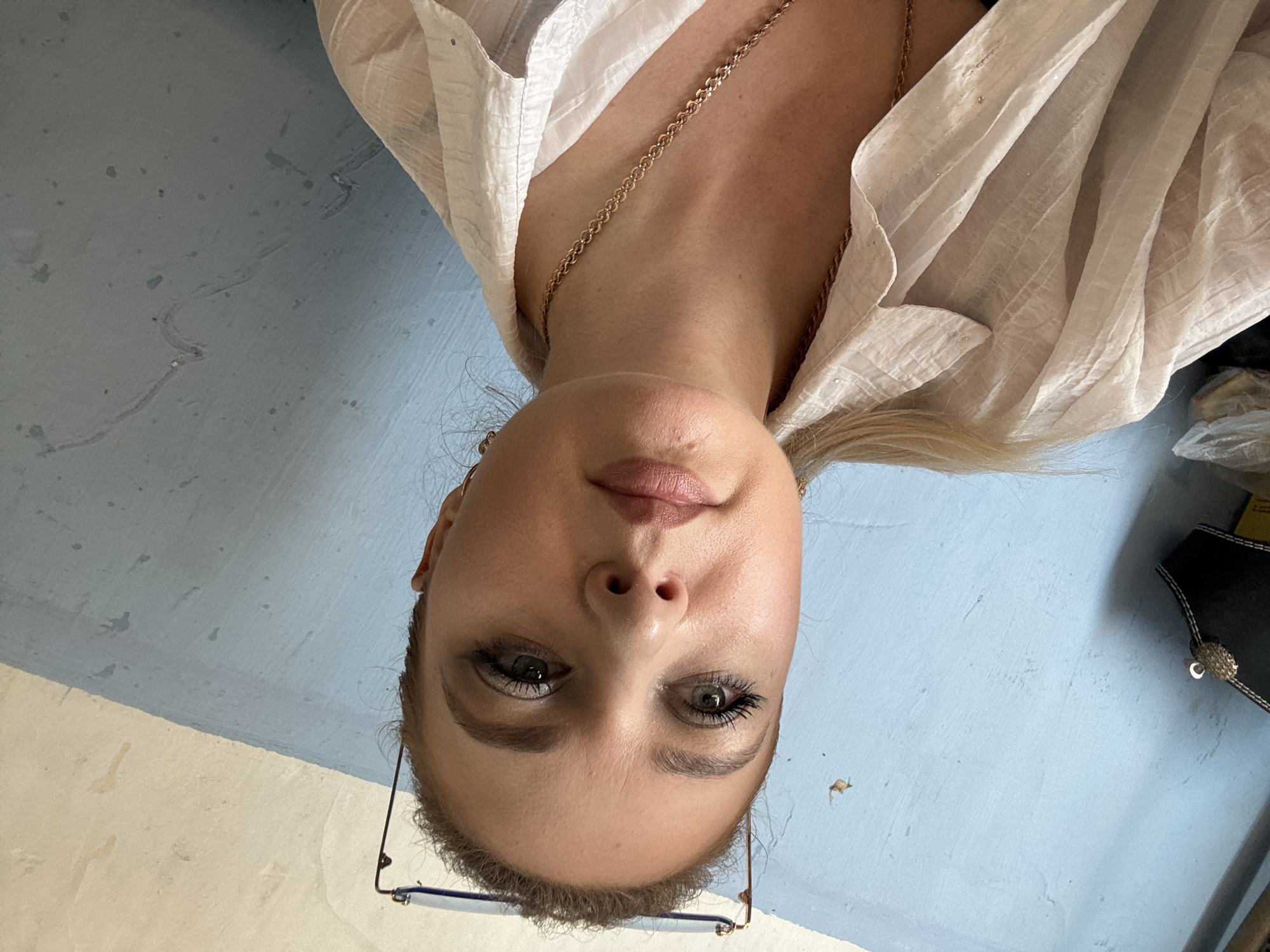 The image is an extreme close-up, upside-down, selfie of a young woman. She has light-colored hair and is wearing a white, sheer, button-up shirt with a delicate gold necklace. Her face is the primary focus, with her eyes looking directly at the camera. She appears to be wearing makeup, with mascara visible on her lashes and a subtle lipstick shade on her lips. Her eyebrows are well-defined. She is also wearing thin-framed glasses that rest on her nose. The background is a muted blue wall with some textured areas and faint marks. To the right of the woman, a black object with white stitching and a silver embellishment is partially visible. The lighting suggests an indoor setting, possibly with natural light, though the time of day is not discernible. The overall emotion conveyed is one of casualness and direct engagement with the viewer.