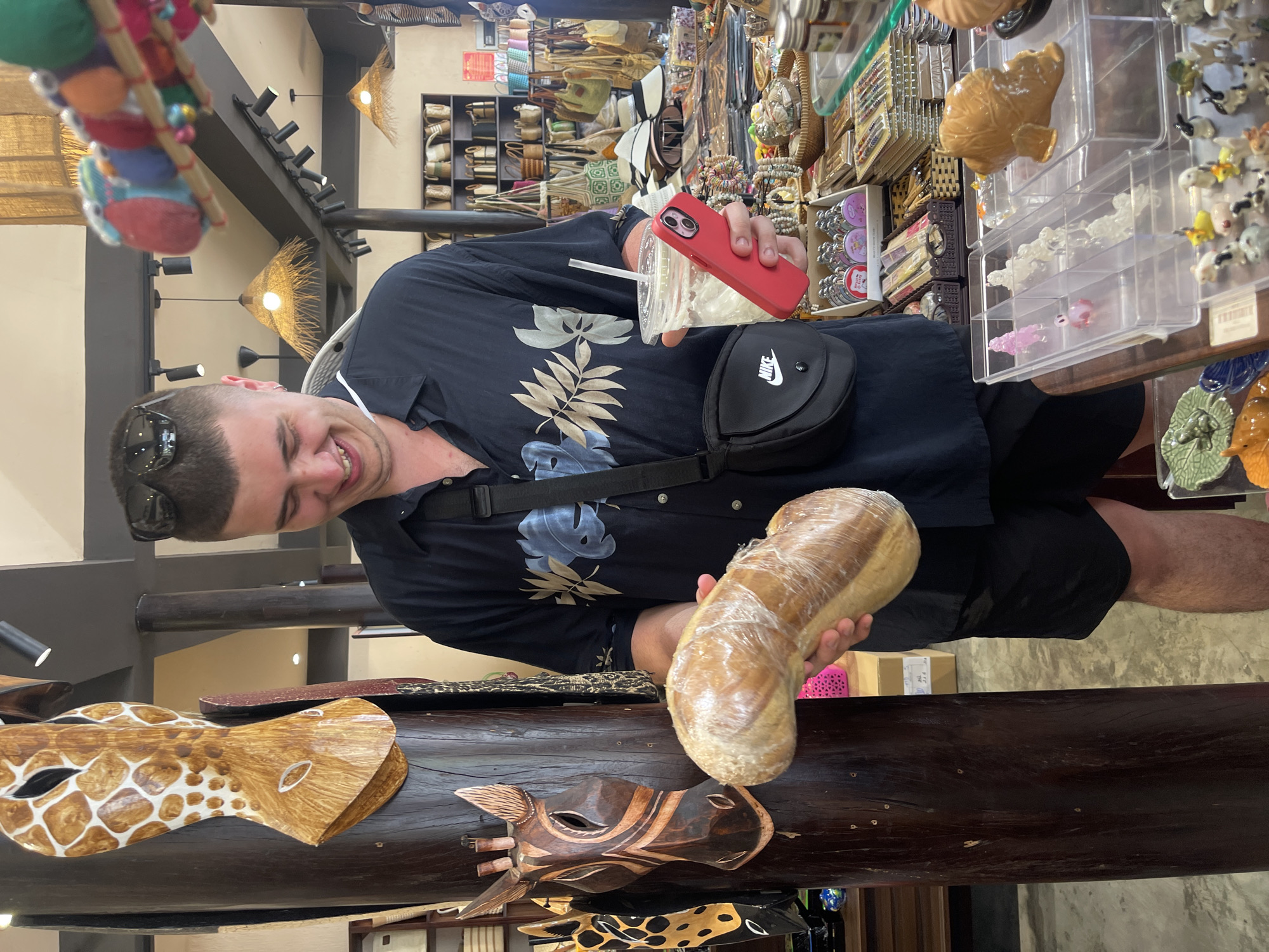 The photo shows a man indoors in a shop in Nha Trang, Vietnam. He is holding a wrapped loaf of bread in his left hand and a drink in a plastic cup in his right hand. He wears sunglasses on top of his head and has a black Nike bag across his torso. He wears a short-sleeved black shirt with a floral pattern and black shorts. He looks to the left and smiles. In front of him is a dark brown wooden table with wooden carved animal masks. Behind him, shop shelves are filled with various souvenirs. Overhead are lights and what appears to be decorative straw ornaments.