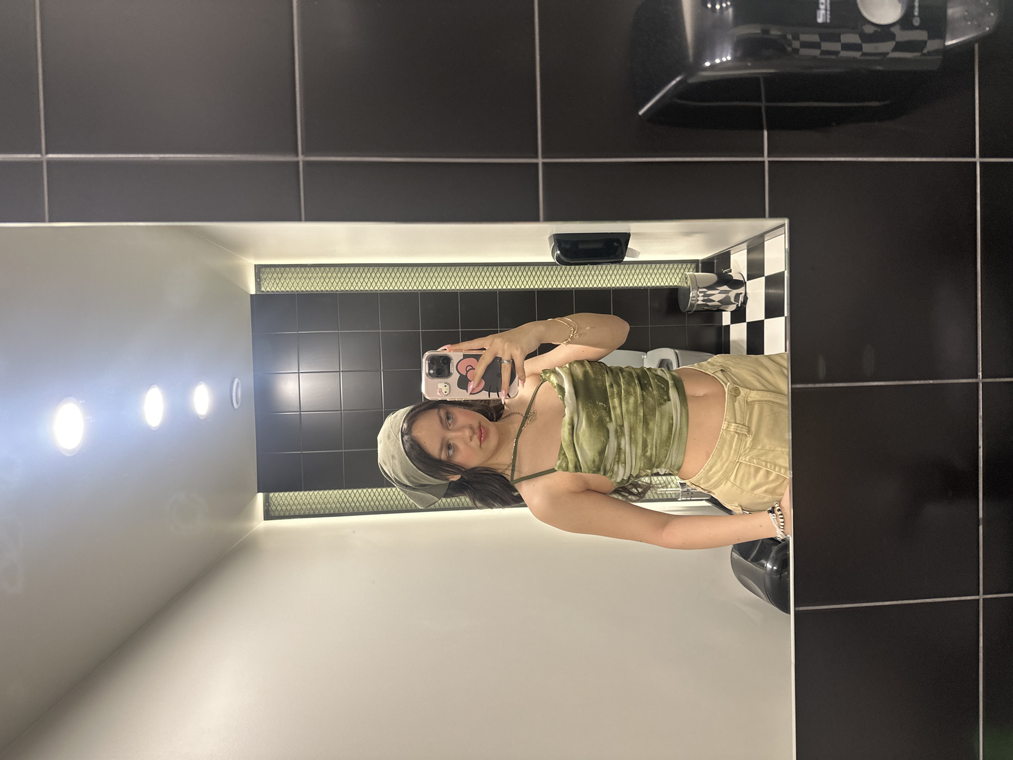 A young woman is taking a selfie in a bathroom mirror. The bathroom features black and white tiled walls, a sink, and overhead lighting. The location is not specified, therefore Yerevan, Armenia cannot be determined from the image.