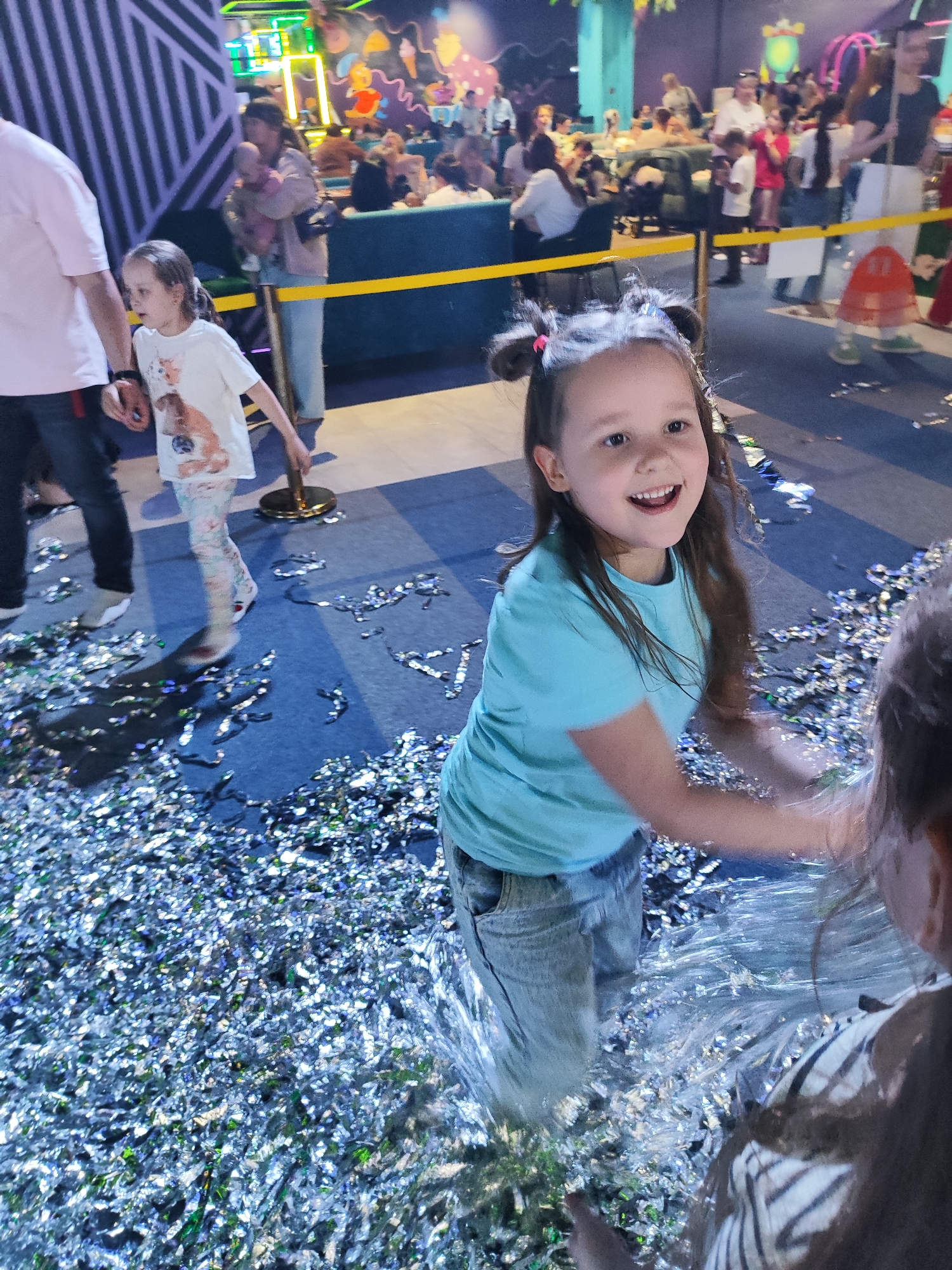 Scene: A brightly lit indoor entertainment venue in Krasnodar, Russia, filled with a crowd of people. The foreground features a child in a light blue t-shirt and jeans, partially surrounded by silver confetti scattered across the floor. The child is smiling and appears to be interacting with another person partially visible in the lower right corner of the frame.
People: In addition to the primary child, there are numerous other people visible in the background. A child in a white t-shirt with a cat image and patterned leggings walks behind a person in a pink shirt. Other adults and children can be seen in a seated area.
Objects: Key objects include the silver confetti littering the floor, tables, chairs, and decorations. A yellow rope line separates the foreground from the background.
Actions: The primary child is engaged in activity, likely playing with the confetti. The child in the white t-shirt appears to be walking. People in the background are seated, possibly eating or watching activities.
Environment: The venue is illuminated with bright overhead lights. The walls are decorated with colorful designs. A seating area is defined by the yellow rope, and other decorations are scattered throughout the space.