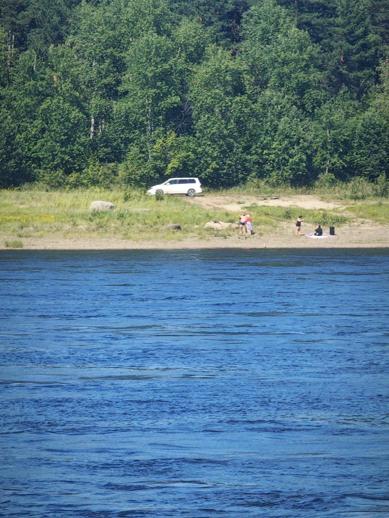 A white van is parked on a grassy area near a sandy riverbank. Several people are visible on the bank. One group appears to be interacting near the van. Further down the bank, a person in dark clothing stands near a mat, and another person sits beside a dark bag. The foreground is dominated by the rippling surface of a blue body of water. The background features a dense forest of green trees.