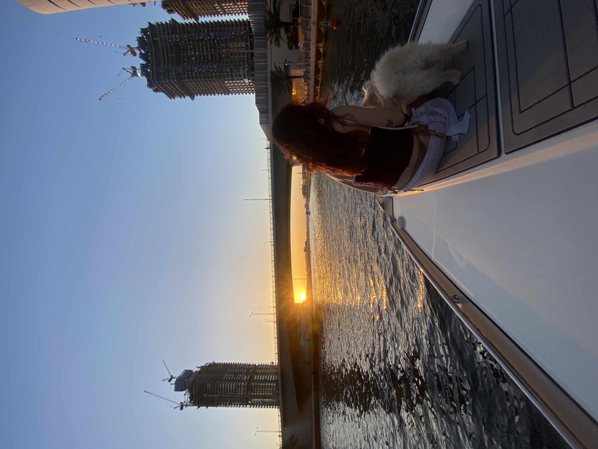 The image captures a scene at sunset on a boat in Dubai, United Arab Emirates. A woman with long, reddish-brown hair is seated on the deck of the boat. A white dog lies next to her. In the background, there is a bridge, and through the bridge, the setting sun is visible. The cityscape features buildings under construction, including skeletal high-rises with cranes. The water is reflective with the colors of the sunset. The boat has a gray deck.
