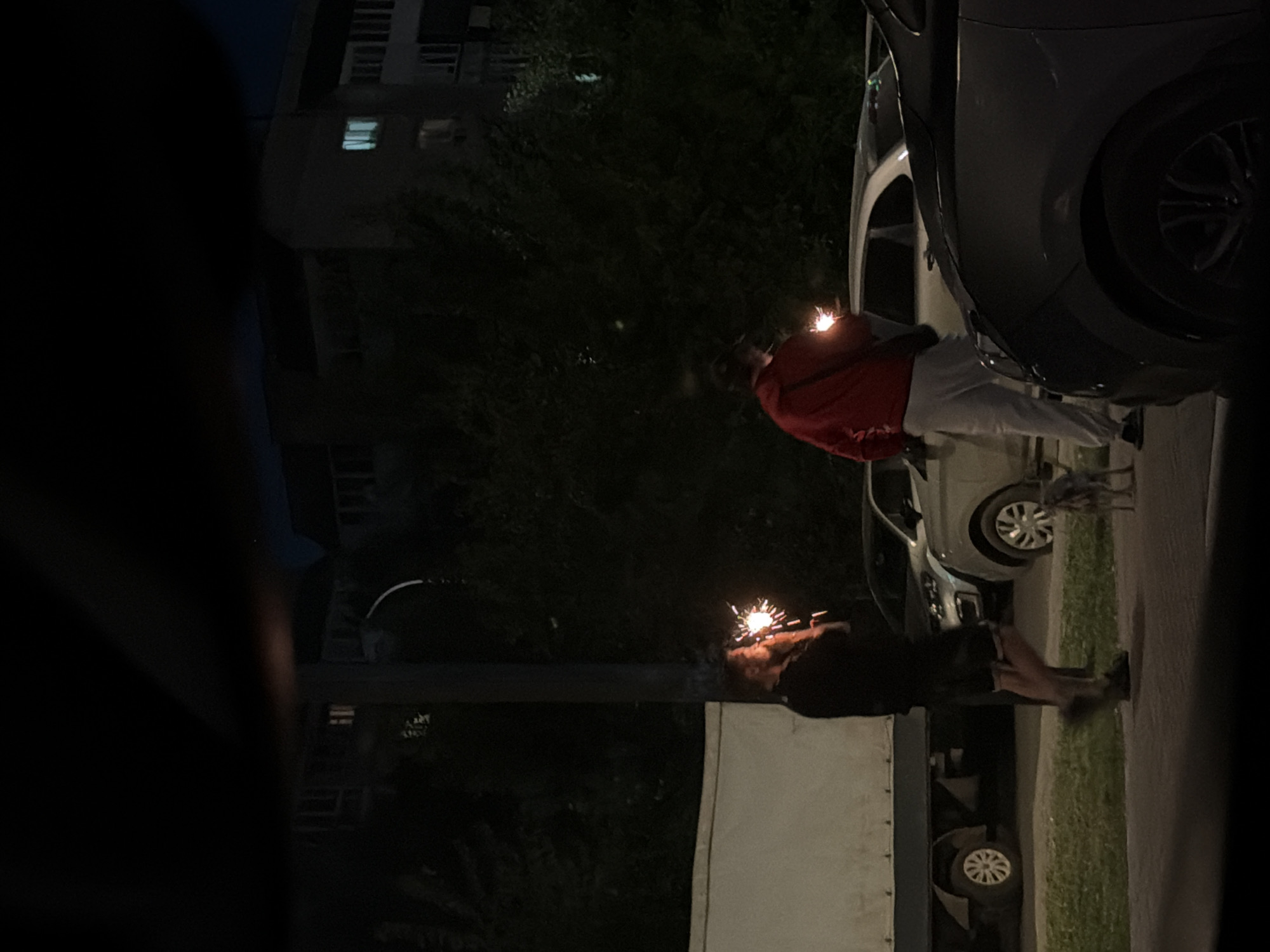 Night shot in Izhevsk, Russia, reveals two people holding lit sparklers outdoors. The image includes portions of parked cars, a building, trees, and a segment of a streetlight. One person is wearing a red jacket and white pants, the other in dark clothing.