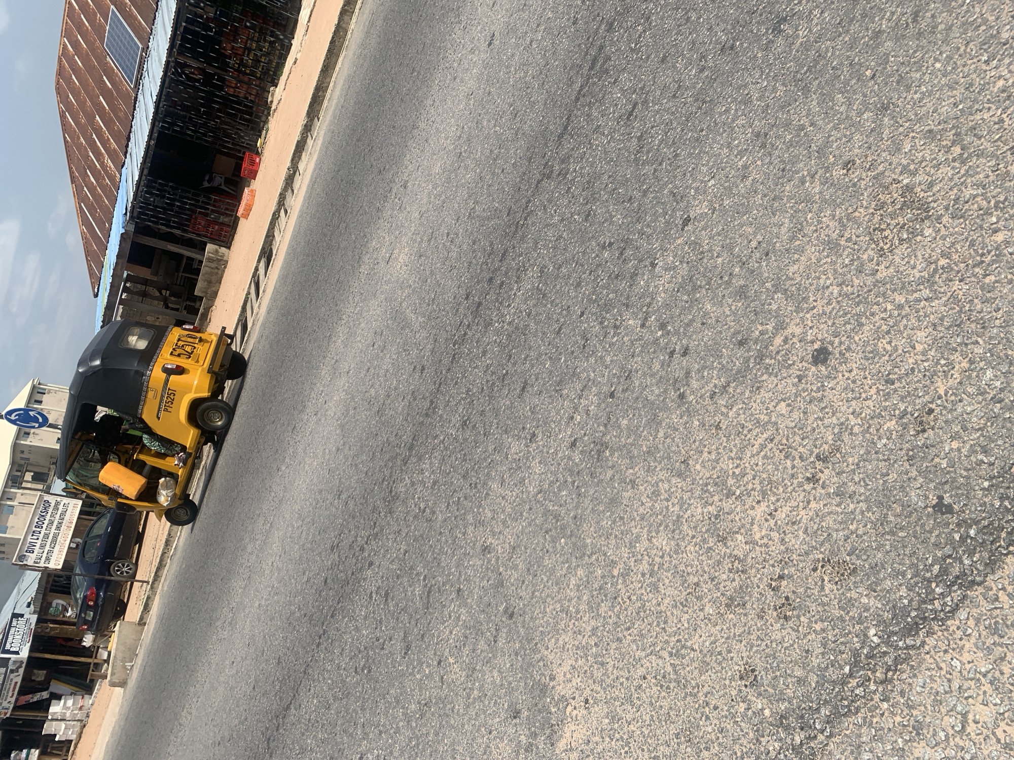 A yellow auto-rickshaw is parked on the side of a road in Jalingo, Nigeria. The auto-rickshaw has a black roof and is carrying passengers. There are several buildings visible in the background, including a bookshop and a shop with a corrugated metal roof. A dark blue car is parked behind the auto-rickshaw. The road is paved and appears to be in good condition. The sky is cloudy.