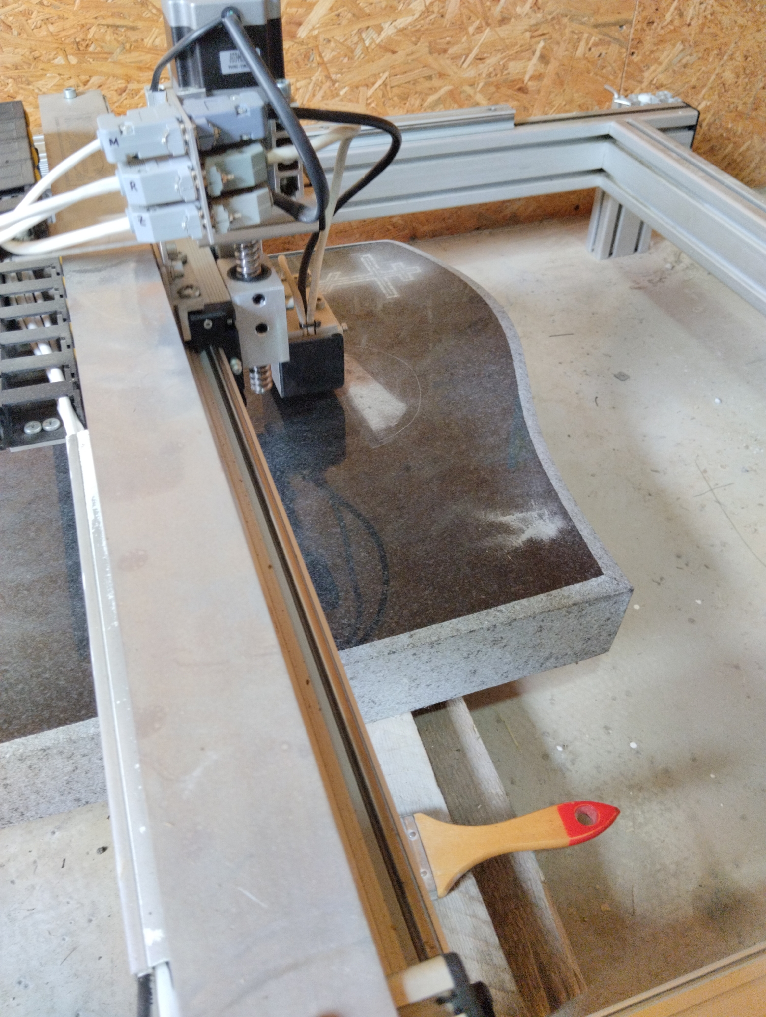 An overhead shot shows a CNC machine engraving a stone slab with a curved edge in an indoor workshop. The machine's engraving head, with three labeled motor drives (M, R, Z), is positioned above the stone. The stone appears to be dark granite and is partially engraved with a cross symbol. A wooden brush with a red plastic handle lies on the floor next to the machine, suggesting recent use or preparation for cleaning. The workshop environment is characterized by wooden walls and a concrete floor, with some debris scattered around. The scene is well-lit, highlighting the details of the machinery and the stone.