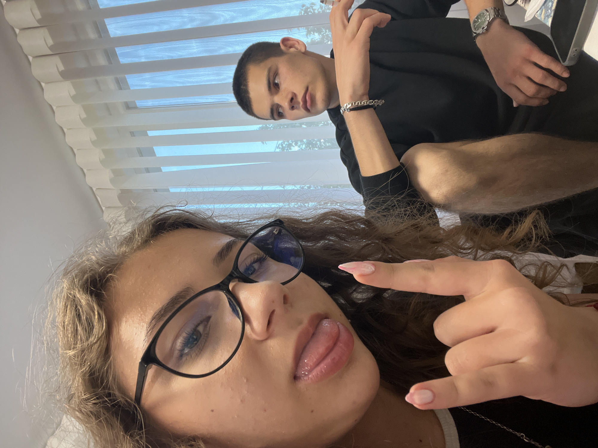 Two individuals are visible in the photograph. A young woman with curly hair and glasses is in the foreground, sticking out her tongue and making a "rock on" hand gesture. In the background is a young man looking towards the camera. He is wearing a black top, a watch on his wrist, and a bracelet. A window with blinds is visible in the background. The specific location in Karavelovo, Bulgaria is not discernible from the image.
