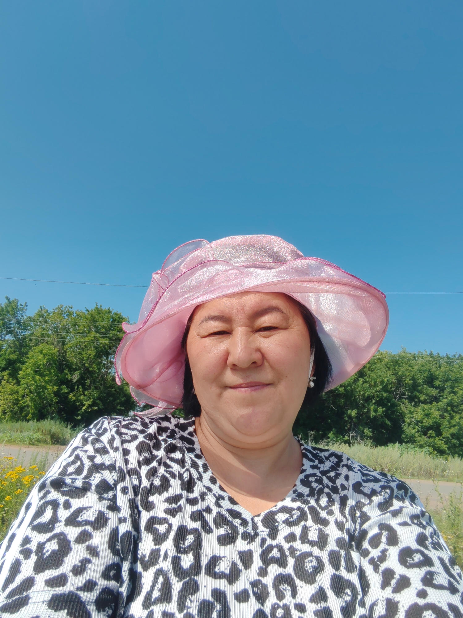 A woman wearing a pink hat and a leopard-print shirt is looking at the camera. She has dark hair and is smiling. The background is a clear blue sky with some green trees and yellow flowers. The image appears to be taken outdoors on a sunny day. The location is not explicitly mentioned but the context of the prompt suggests it is in the city of Акжар, Kazakhstan.