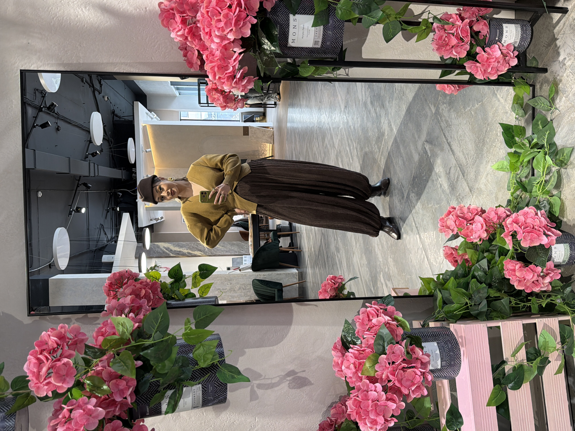 This image, rotated from its original orientation, captures a full-body mirror selfie taken indoors. A person, dressed in a stylish outfit, stands in front of a large, black-framed mirror, holding a light green smartphone to take the picture. They are wearing a dark brown beret-like hat, a mustard yellow or olive green V-neck sweater, loose-fitting pleated brown trousers, and black boots. Gold-colored earrings and rings accessorize their look.
The mirror's frame is heavily adorned with artificial floral and green decorations. Vibrant pink hydrangeas and lush green leafy vines cascade around the edges, originating from dark grey patterned containers. Text visible on these containers includes "MONS Acrylic Paint" and "CASHMERE," suggesting the presence of decorative items or products related to home decor or art supplies. A partially visible light pink wooden slatted box or crate is also present on the right side of the mirror's base.
The reflection in the mirror reveals a modern and well-appointed interior space. The floor is covered with light grey tiles arranged in a diagonal pattern. The background within the reflection shows dark grey or black walls illuminated by several white circular lights and spotlights. Further back, white counters or shelving and dark green upholstered chairs are visible, indicating a sophisticated showroom, boutique, or office environment. The scene appears to be set during the daytime, illuminated by both artificial and possibly some natural light. The person's expression is neutral, focused on capturing the mirror shot.