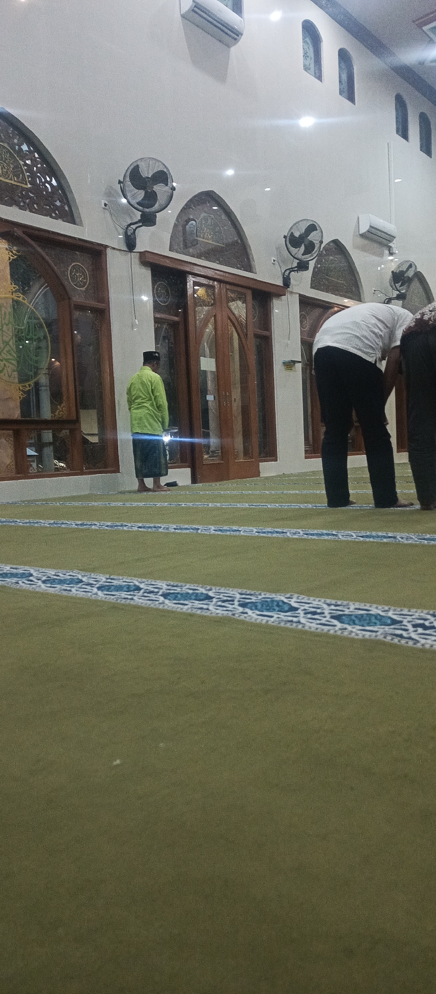The image depicts the interior of a mosque, likely located in Arjasa, Indonesia, during prayer. The floor is covered with a green carpet featuring a blue and white geometric pattern. In the foreground, the carpet extends towards the viewer, with rows of prayer mats visible in the midground.
There are at least three individuals present. One person, wearing a lime green long-sleeved shirt and a sarong, stands facing the wall in the midground, appearing to be in the middle of prayer or awaiting the start of prayer. Another individual, dressed in a white t-shirt and dark pants, is bending over in the right midground, also seemingly engaged in prayer. A third person, partially visible on the far right, is also bent over.
The background shows the mosque's architecture, including large wooden-framed windows with decorative elements and stained glass inserts. Wall-mounted fans are visible on the upper walls, and air conditioning units are mounted high on the walls. The lighting appears to be a mix of artificial light and possibly some natural light filtering through the windows. The overall atmosphere suggests a place of worship.