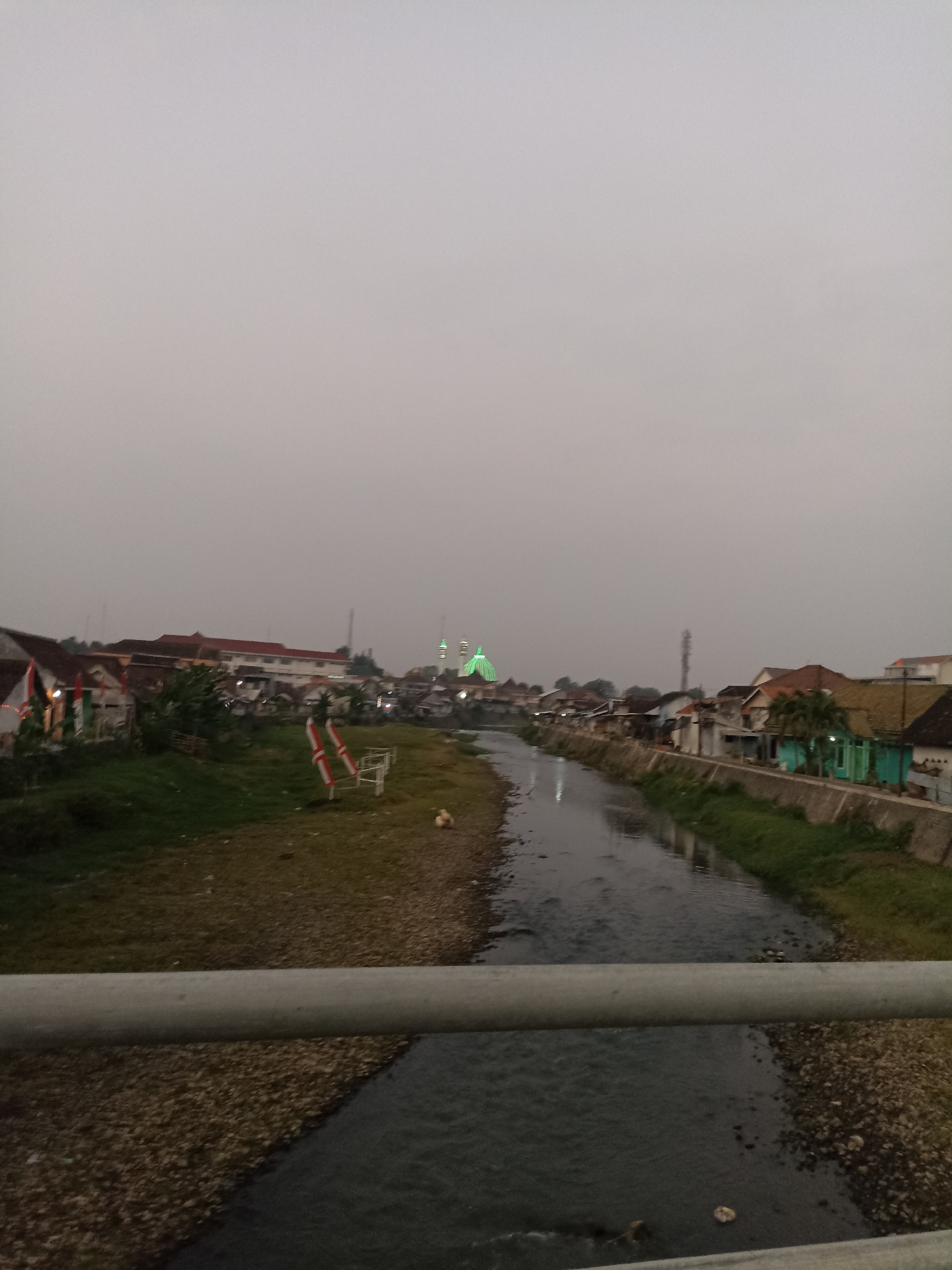 A wide shot captures a tranquil river flowing through a populated area, likely Lumajang, Indonesia, under an overcast sky. The river, appearing somewhat murky, is bordered by a grassy embankment on the left and a concrete retaining wall on the right, lined with houses. A prominent railing runs across the foreground of the image, suggesting the photo was taken from a bridge or elevated walkway. On the left bank, a few small buildings with red-tiled roofs are visible, with Indonesian flags adorning some of them. In the distance, a mosque with green illuminated domes stands out against the hazy horizon, indicating its significance within the community. The houses on the right bank exhibit a variety of architectural styles and colors, with some appearing older and more weathered than others. The overall atmosphere is serene and subdued, possibly indicating dusk or dawn.