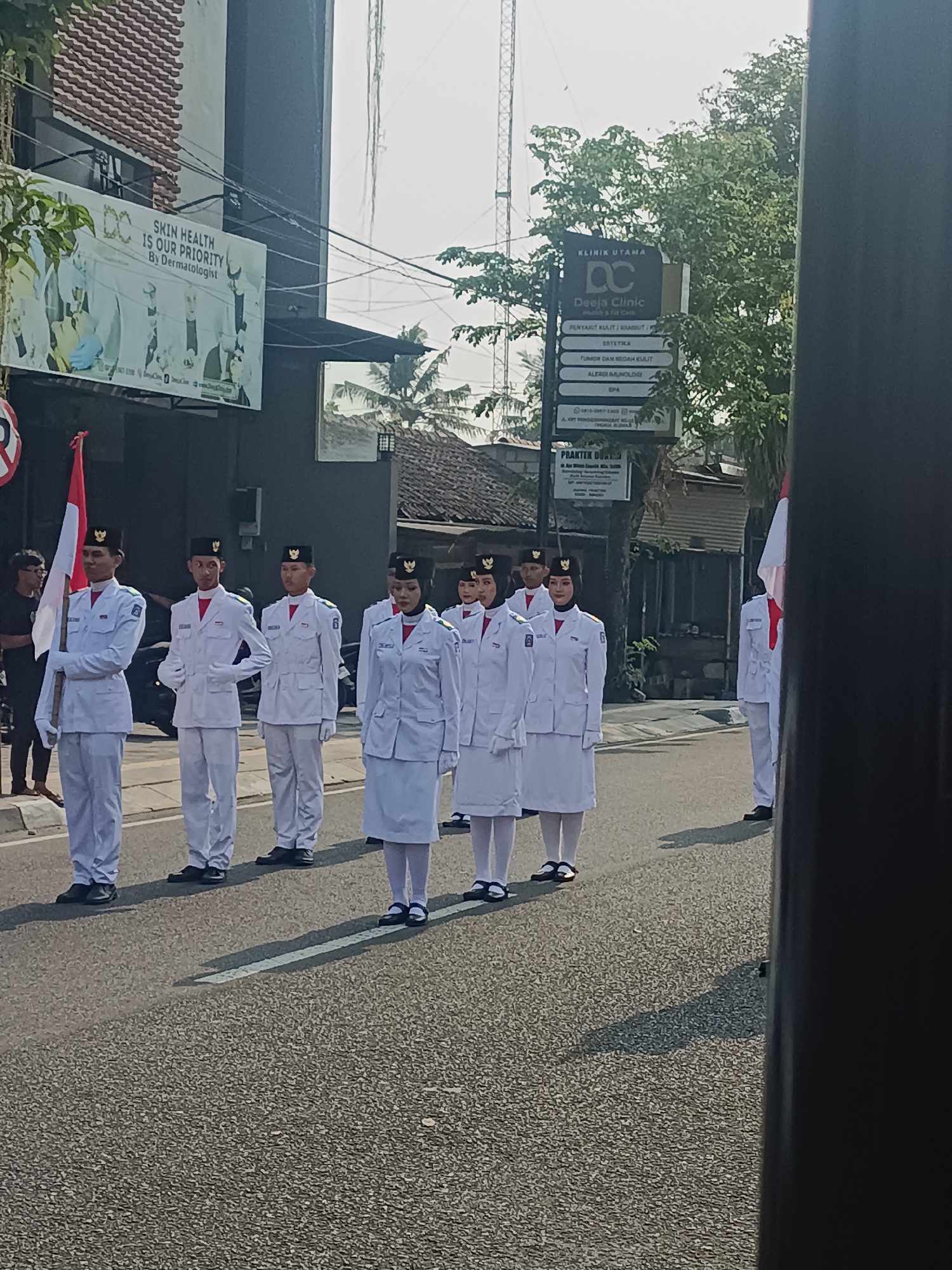 In the city of Sleman, Indonesia, a group of individuals in white uniforms with black hats stand in formation on a street. They appear to be a marching band or a ceremonial group, possibly a school group. One individual holds a red and white flag. Behind them, buildings line the street, including a clinic with a sign that reads "KLINIK UTAMA DC Deeja Clinic" and "PRAKTEK DOKTER." The atmosphere is bright, suggesting it is daytime. The individuals are of various ages and genders.