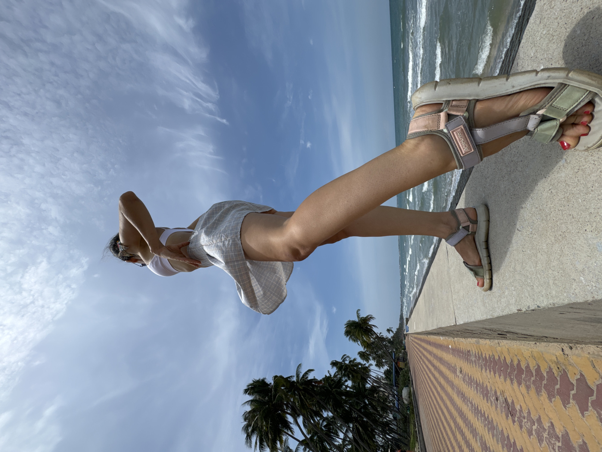 A low-angle, full-shot photograph captures a person standing on a concrete ledge overlooking the sea in Phu Thuy Ward, Vietnam. The individual, with their back to the viewer, wears a white bikini top and a light-colored, plaid skirt that billows in the wind. Their right arm is bent upwards, touching their hair, while their left hand rests on their hip. They wear light-colored sandals with pink and gray accents. The scene is set against a backdrop of a vibrant blue sky with wispy white clouds. To the left, a row of palm trees lines a patterned walkway with yellow and red brick pavers. The turquoise sea stretches out to the horizon, with waves gently lapping against the shore. The image conveys a sense of breezy, coastal relaxation.